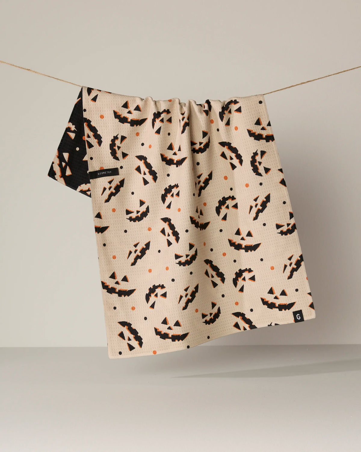 The Geometry “Haunted Grin” tea towel, crafted from recycled materials, features a beige double-sided design adorned with black and orange jack-o-lantern faces and playful dots, shown hanging on a clothesline against a simple light background.