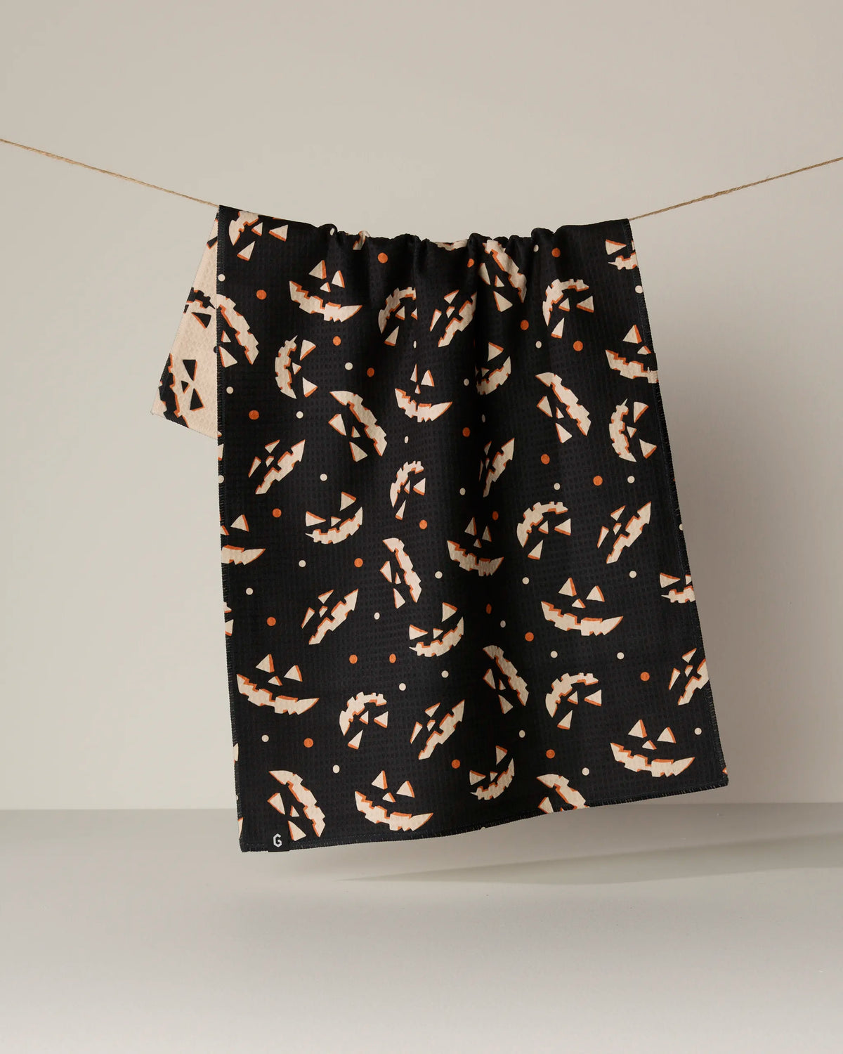 The “Haunted Grin” fabric by Geometry, featuring bold beige and orange abstract patterns on black, hangs on a string against a plain background and is crafted from recycled materials for an eco-friendly touch.