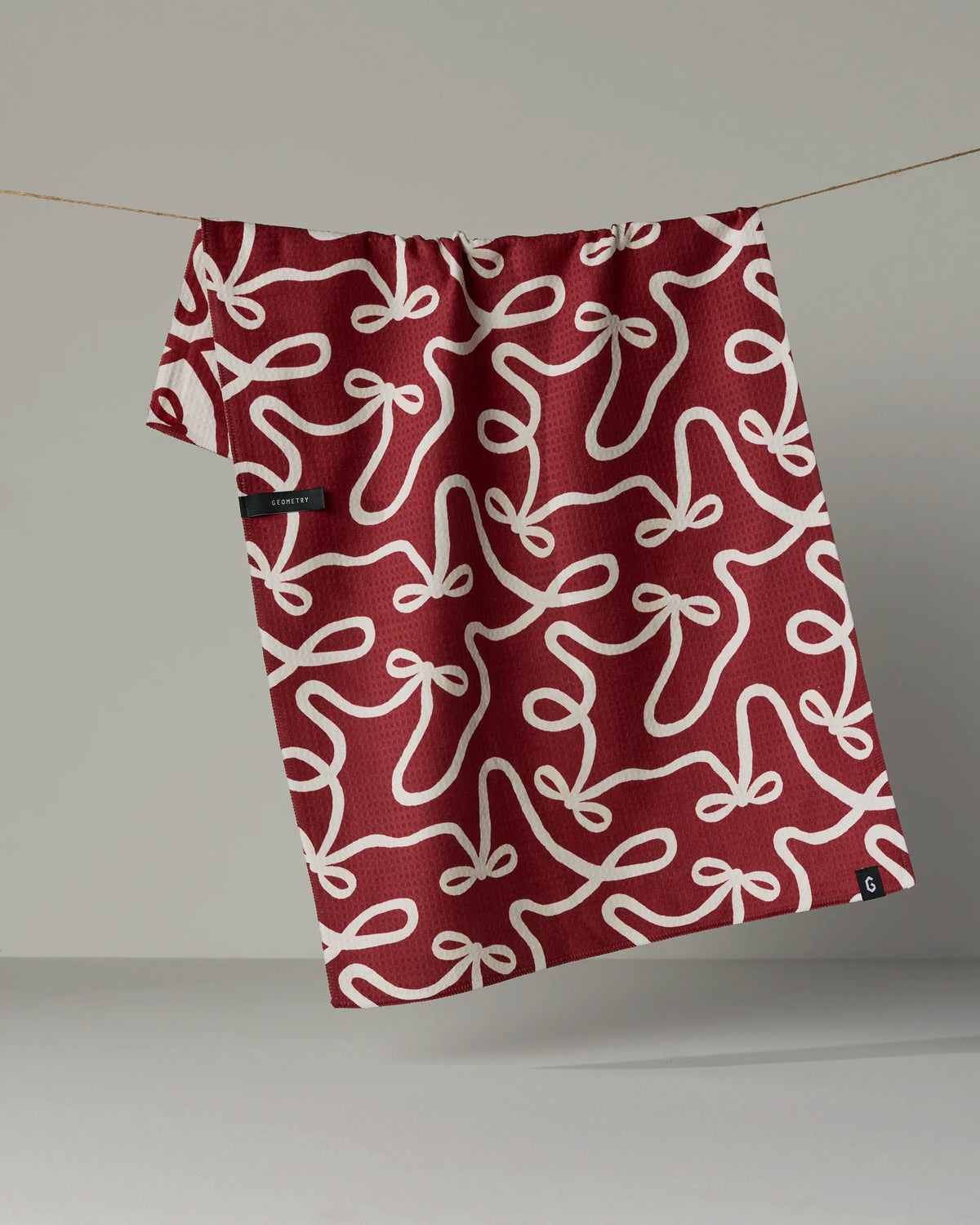 The Geometry Holiday Bows recycled kitchen towel, featuring a white bow pattern on a vibrant red background, hangs on a string against a neutral backdrop.