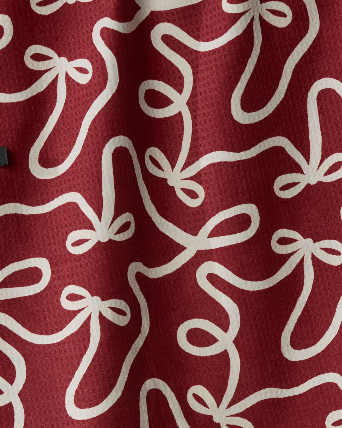 The “Holiday Bows” tea towel by Geometry features red fabric adorned with a textured surface and a repeating pattern of white, loosely sketched bow shapes and looping lines. Designed as a double-sided towel, it drapes softly—its absorbent, quick-dry material ideal for adding both style and function to your kitchen.