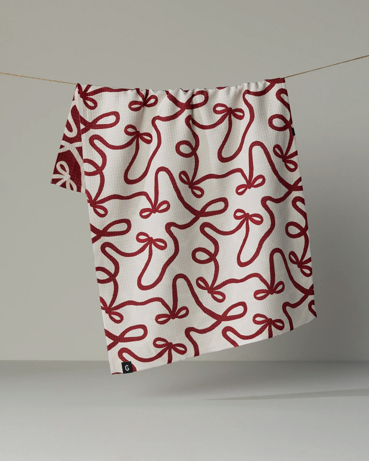 The Geometry Holiday Bows tea towel, featuring a red abstract bow pattern on both sides of crisp white fabric, hangs from a thin string against a plain gray background.