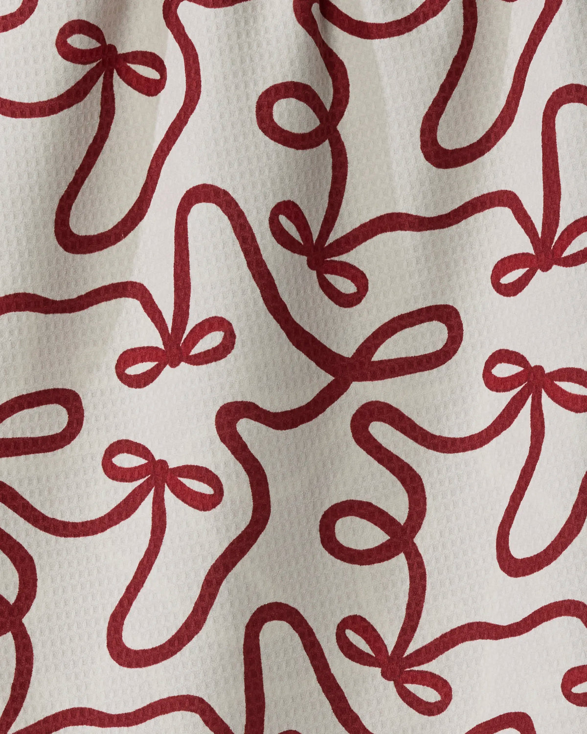 Red bow shapes of varying sizes and orientations create a playful, abstract pattern across the white textured fabric of Geometry’s Holiday Bows towel. The absorbent, quick-dry material—made from recycled fibers—combines practicality with stylish design, while gentle fabric folds add shadow and depth to the lively motif.