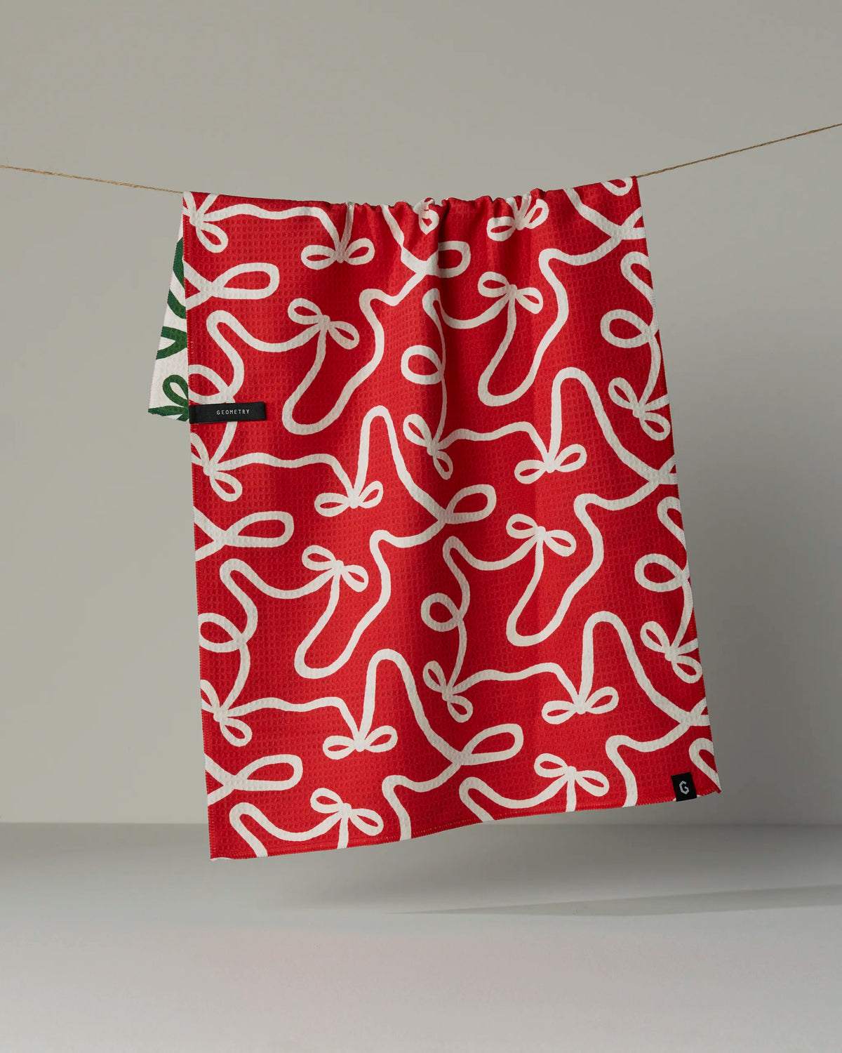 A “Holiday Bows” kitchen towel by Geometry, adorned with a white bow pattern on red fabric and a small green tag on its left side, hangs on a line against a plain gray background.