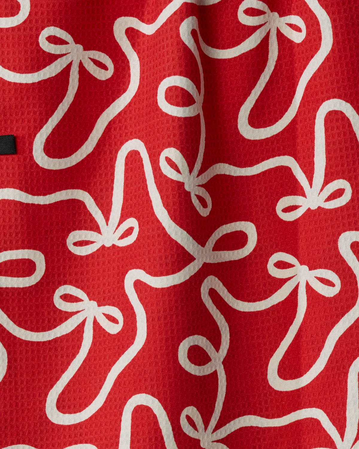 The “Holiday Bows” double-sided tea towel by Geometry features a red textured fabric adorned with a repeating pattern of white shoelace bows, making it a stylish and practical addition to any kitchen.