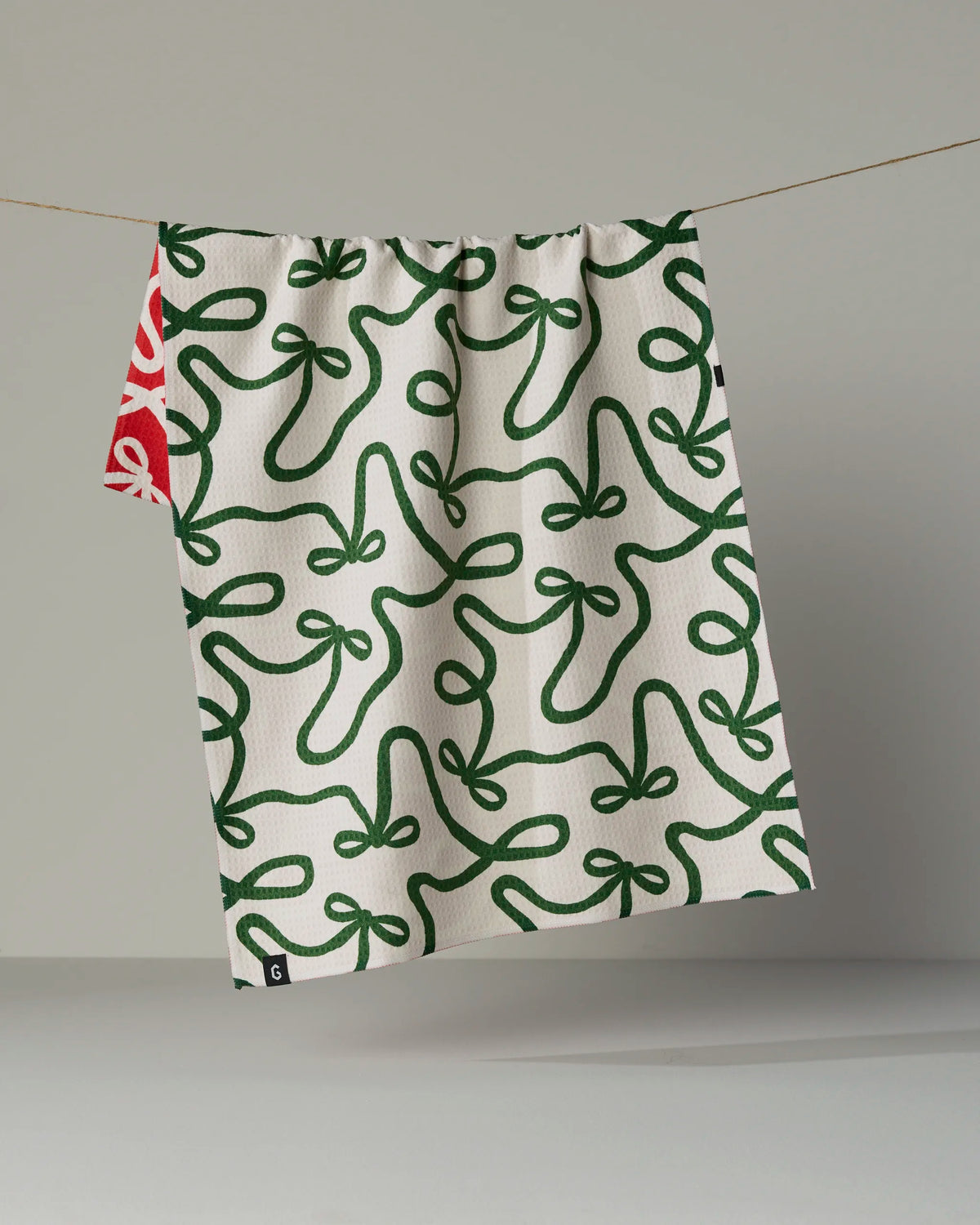 A white, double-sided tea towel from Geometry, featuring the “Holiday Bows” green ribbon pattern, hangs on a line. A small section of a red towel with a white design is visible behind it.