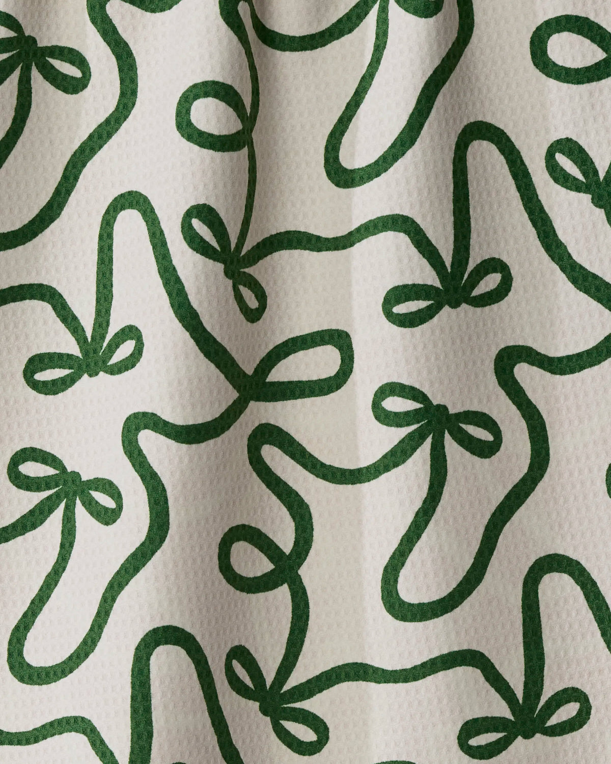 Geometry’s Holiday Bows towel features green ribbon-like loops forming bow shapes on a textured white cloth background with visible fabric folds—an absorbent, quick-dry towel that blends festive design with functional style.