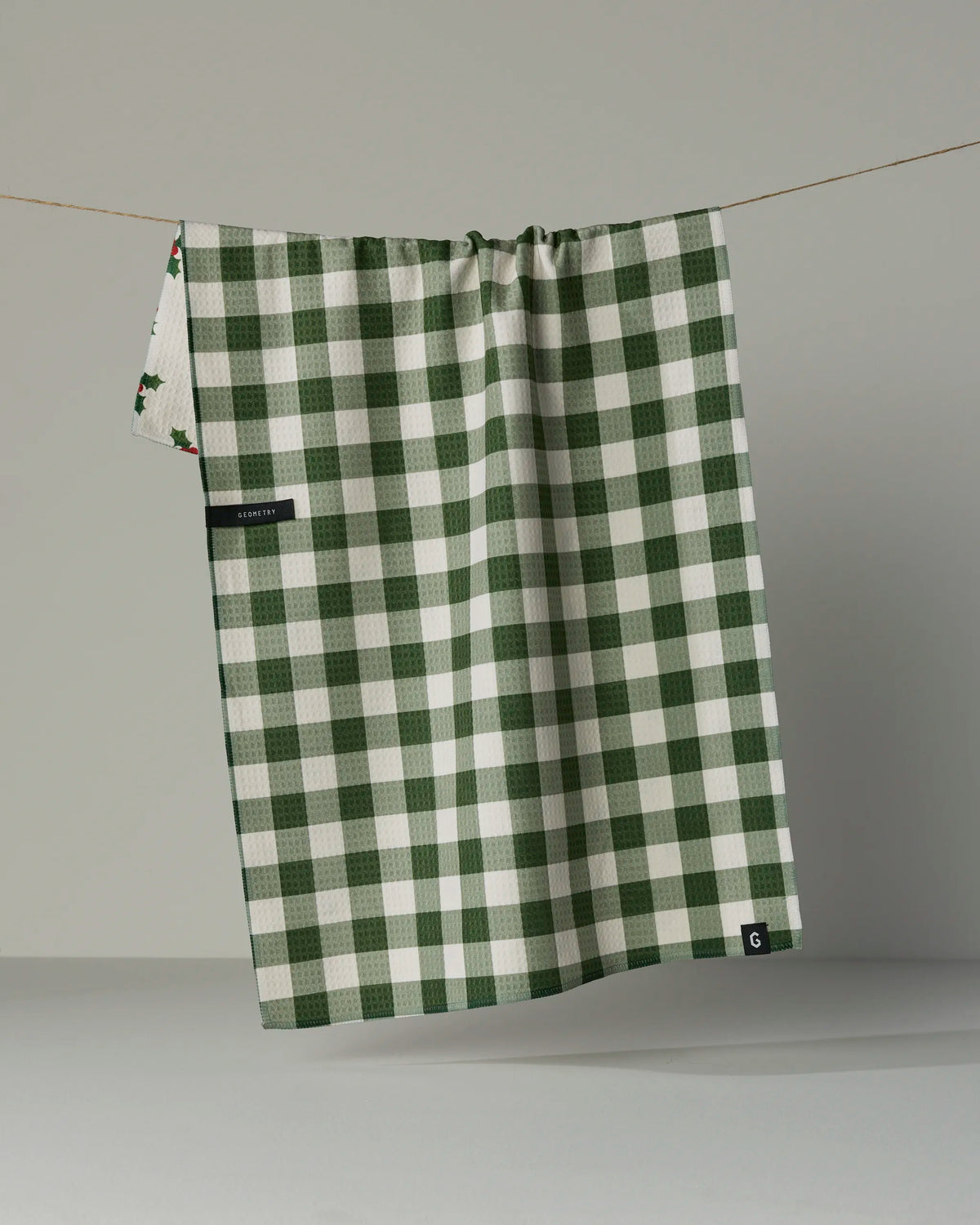 A “Holiday Holly” kitchen towel by Geometry, featuring a green and white checkered pattern and crafted from absorbent, quick-drying recycled materials, hangs on a string against a plain gray background. A small tag and the towel’s decorative double-sided edge are also visible.