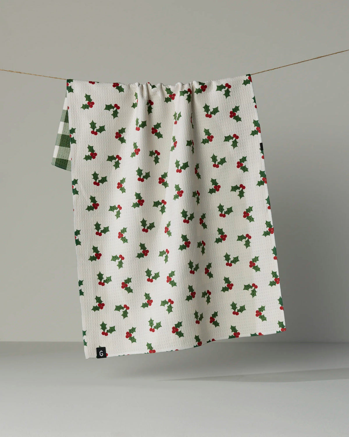 The Geometry Holiday Holly dish towel, crafted from recycled materials and featuring a festive pattern of green and red holly leaves, hangs on a clothesline against a plain gray background.