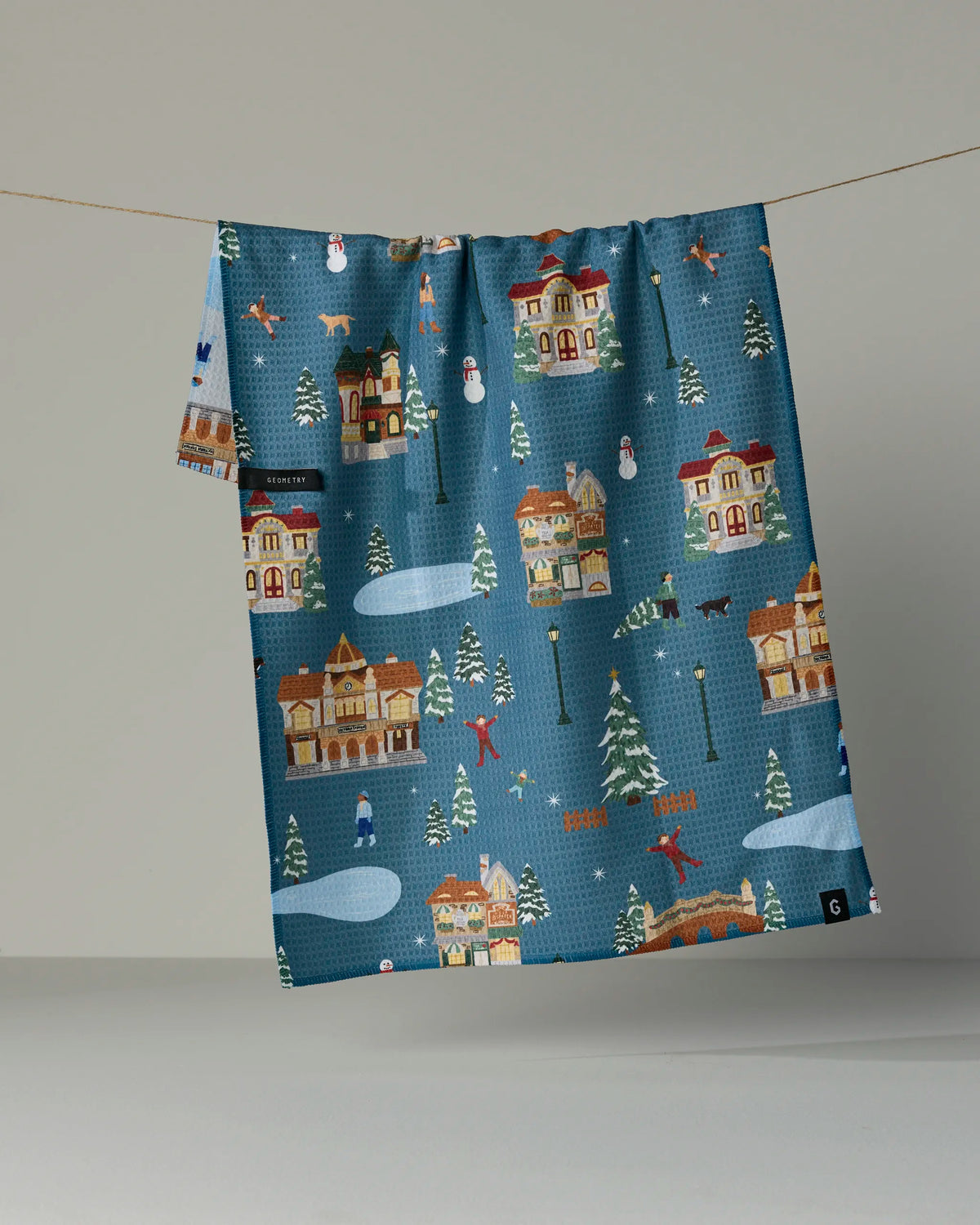 Holiday Nostalgia by Geometry showcases a charming blue fabric tea towel adorned with a festive winter scene—including houses, ice skaters, snowmen, trees, and animals—hanging on a clothesline against a plain background. This delightful double-sided towel is not just visually appealing but also highly absorbent, making it the perfect choice for adding holiday cheer to your kitchen.