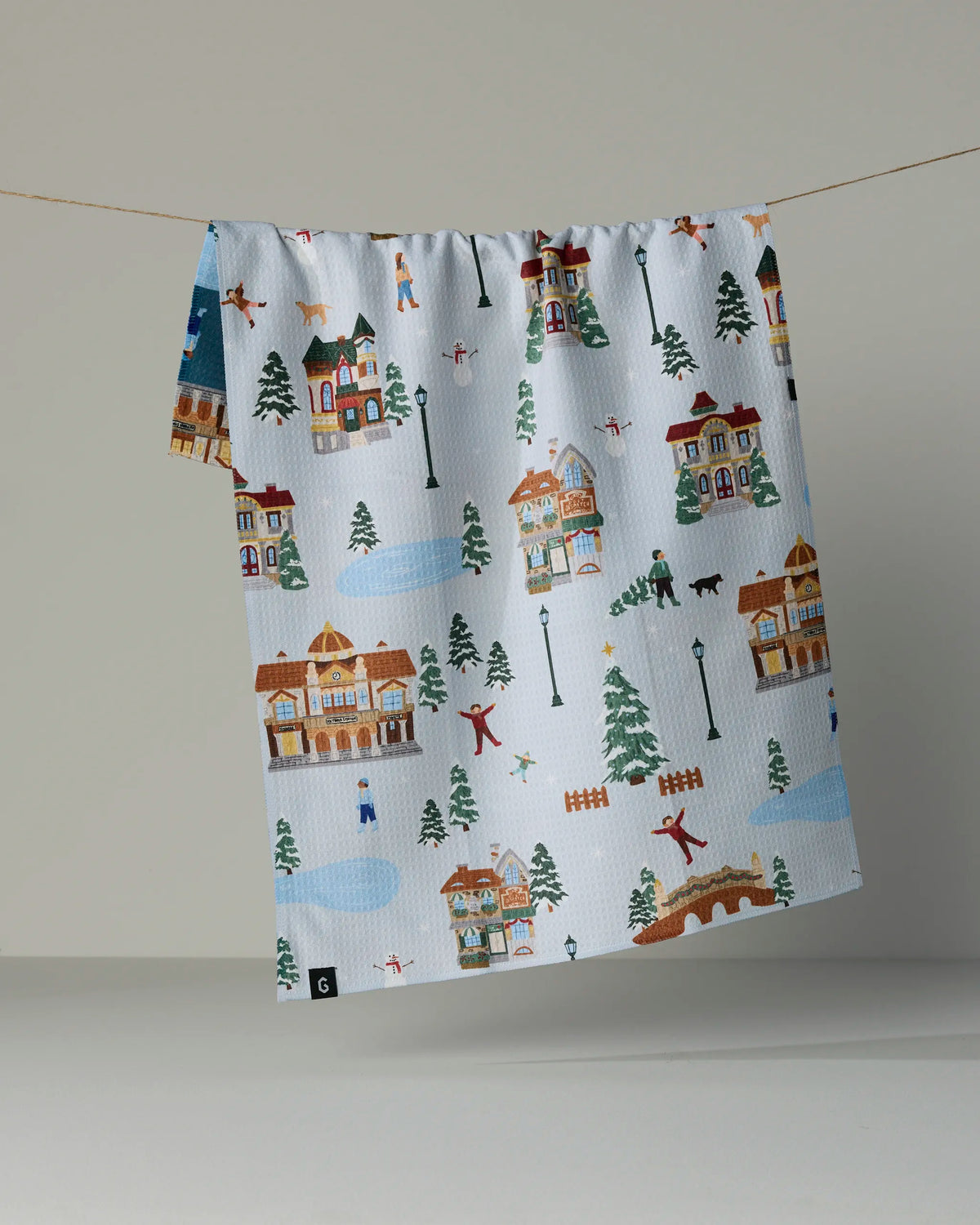 The Geometry “Holiday Nostalgia” double-sided tea towel, featuring a charming winter village scene with houses, trees, people skating, skiing, and walking dogs, hangs on a clothesline against a plain background.