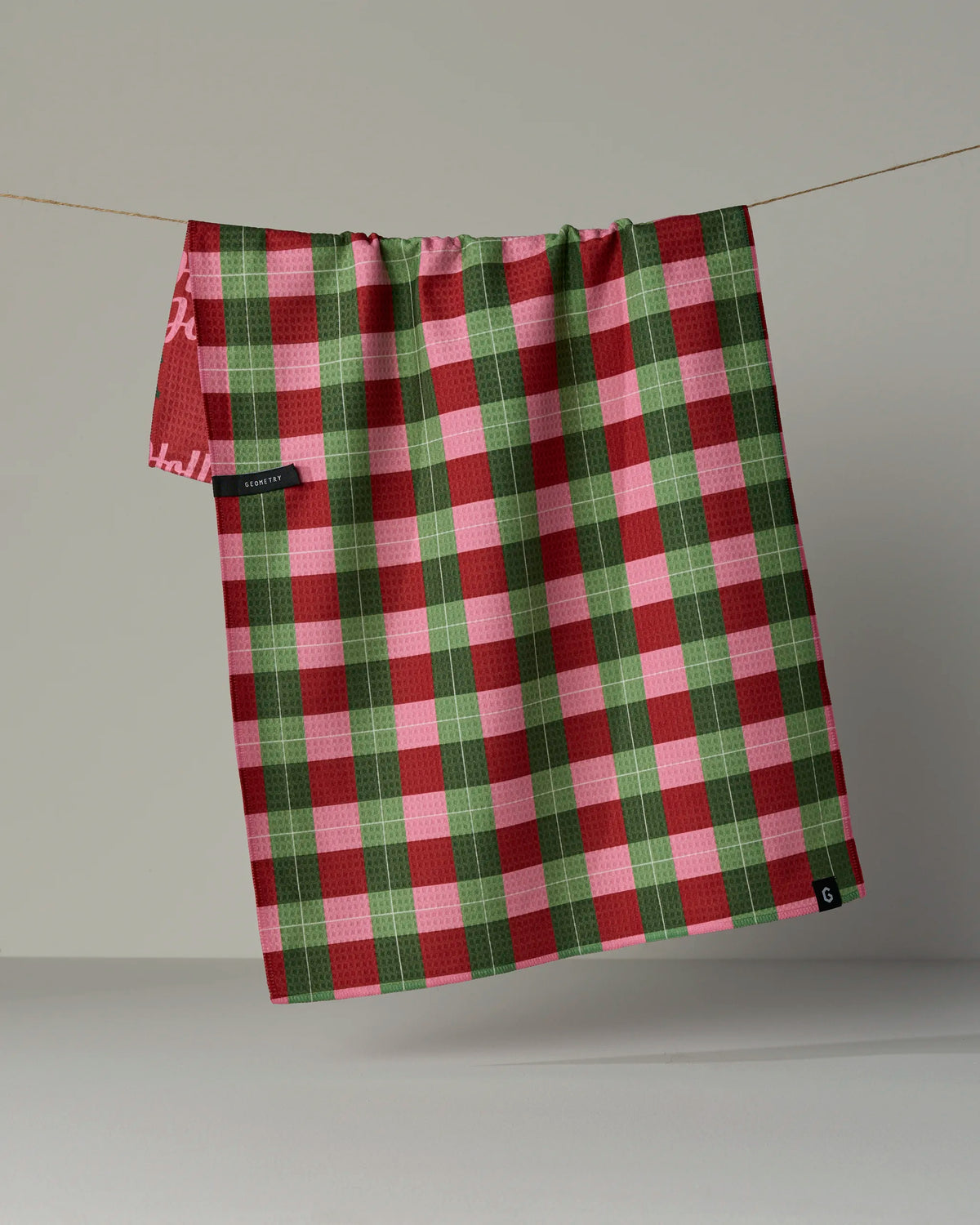The “Holly Jolly Xmas” tea towel by Geometry features a festive red, green, and white plaid design and hangs from a thin string against a neutral background. This double-sided towel includes two black tags—one attached to the bottom edge and another to the side of the fabric.