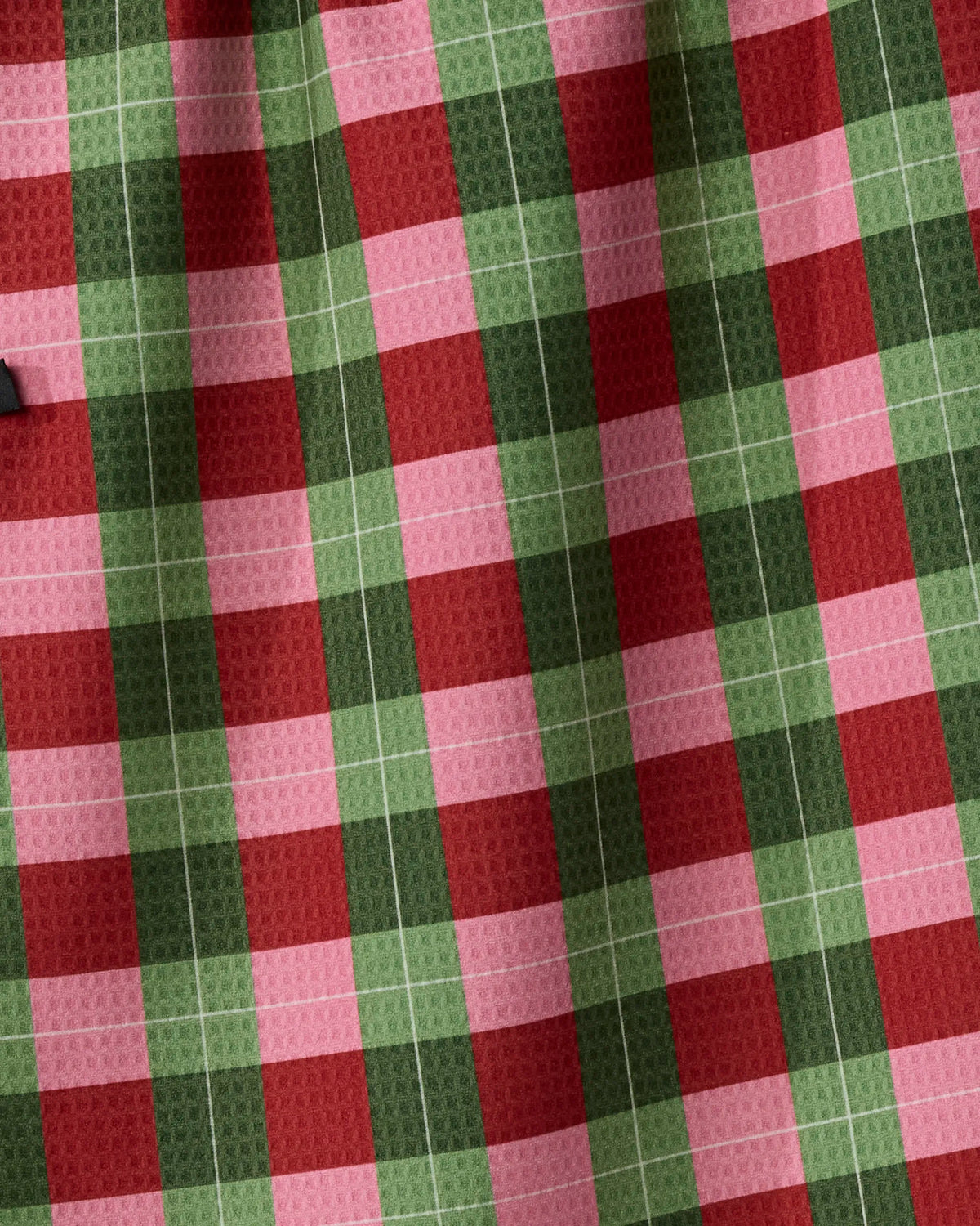 A close-up of the Geometry “Holly Jolly Xmas” kitchen towel showcases its absorbent fabric, adorned with a checkered pattern in red, green, and pink squares. Made from recycled materials, the towel’s slightly wrinkled texture creates gentle shadows and adds visual depth.