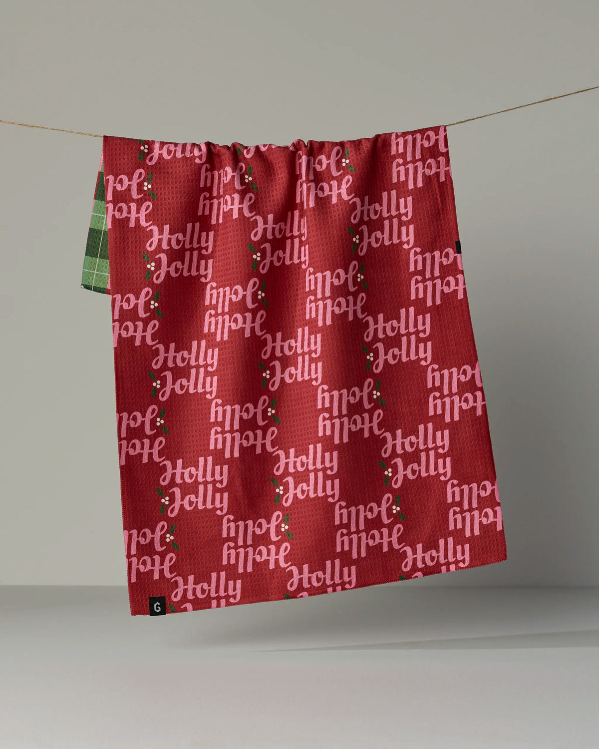 The Geometry Holly Jolly Xmas tea towel is a festive red holiday towel made from absorbent recycled materials, featuring “Holly Jolly” and holly berries printed in white and pink text. This double-sided towel hangs on a line, showcasing a green plaid corner on the back.