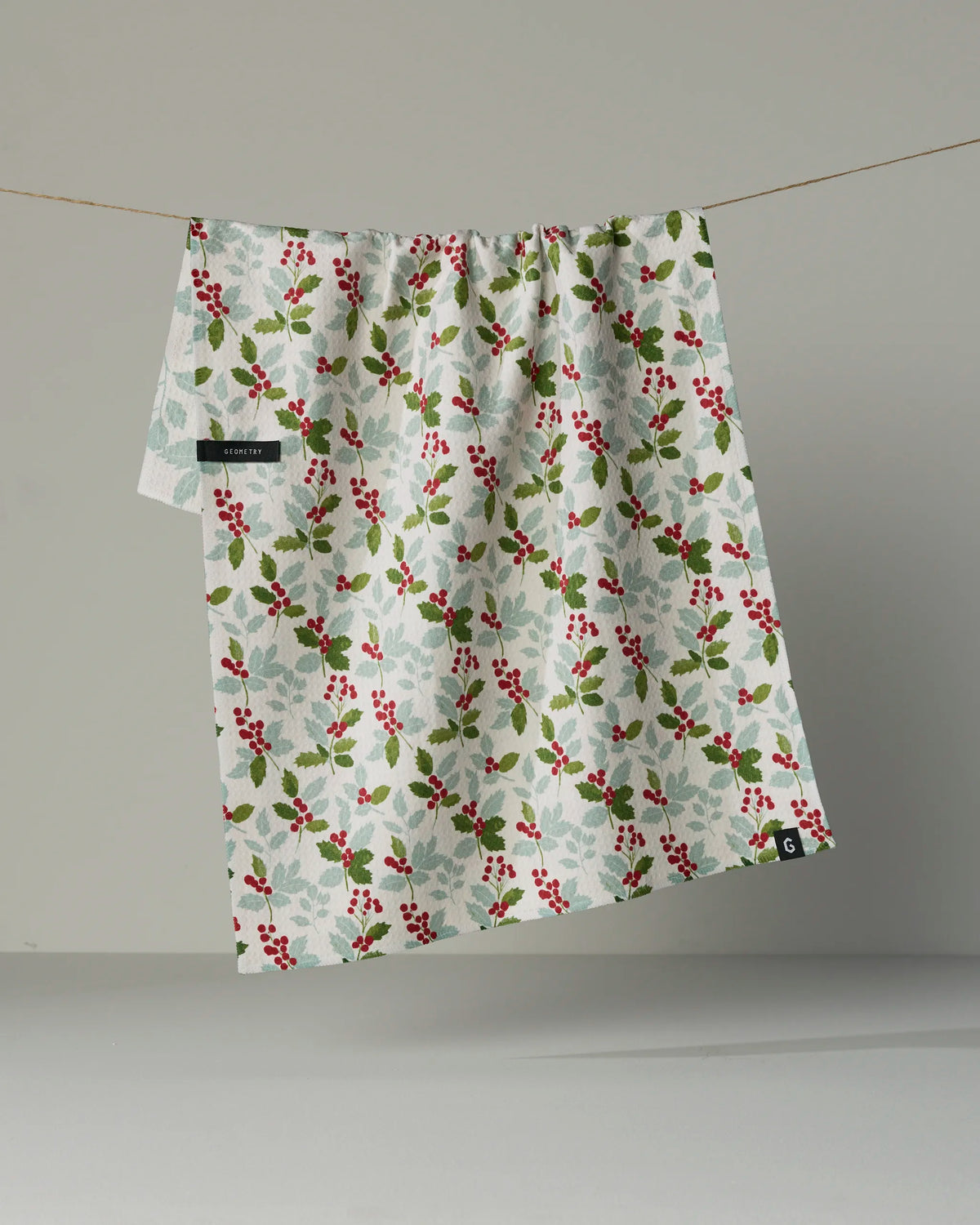 The Geometry “Holly Lane” double-sided tea towel, featuring a festive pattern of green holly leaves and red berries, hangs on a line against a plain background.