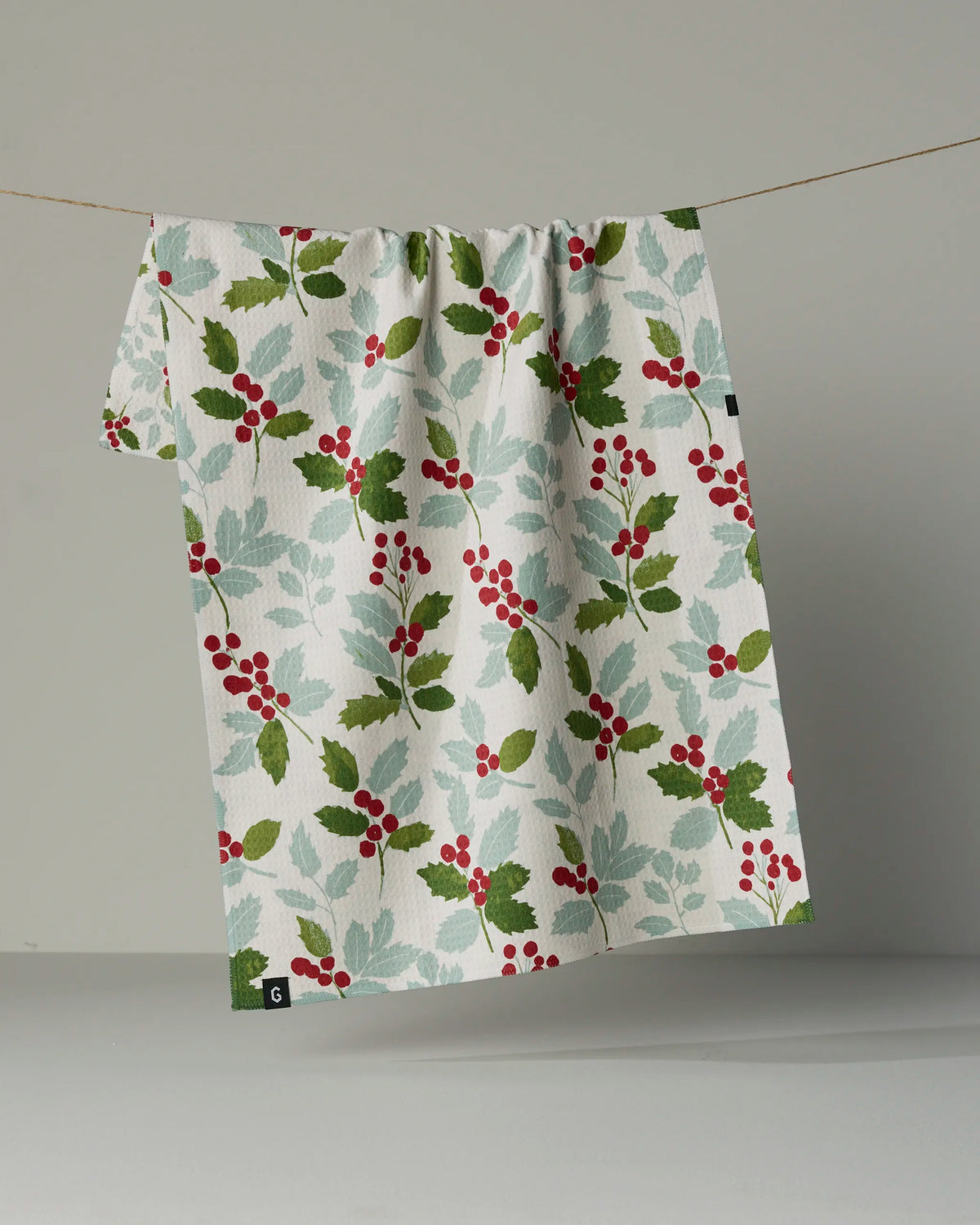 The Holly Lane double-sided tea towel by Geometry, crafted from recycled materials and showcasing a white cloth adorned with a red berry and green leaf pattern, hangs on a string against a plain light gray background.