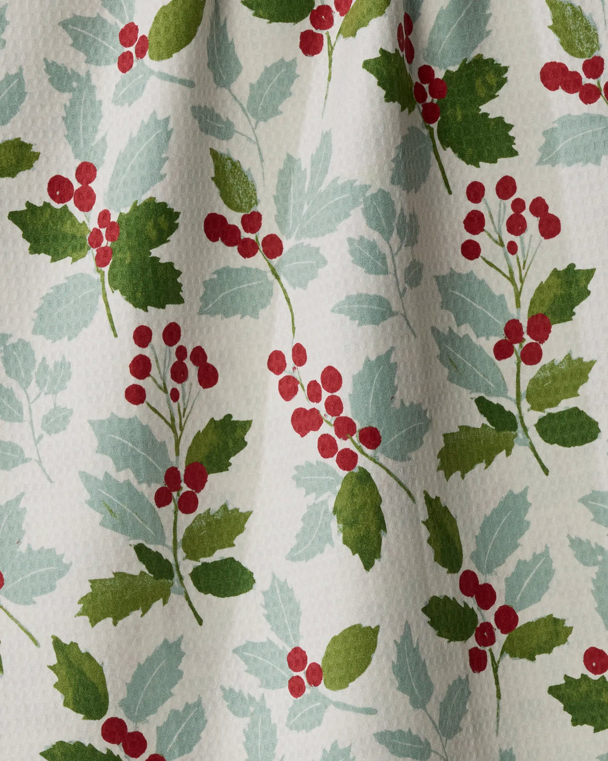 Introducing Holly Lane by Geometry—a quick-drying textile made from recycled materials, adorned with a festive holly pattern of green leaves and red berry clusters on a white background. Light shadows across the fabric hint at soft folds, adding texture to its holiday-inspired design.