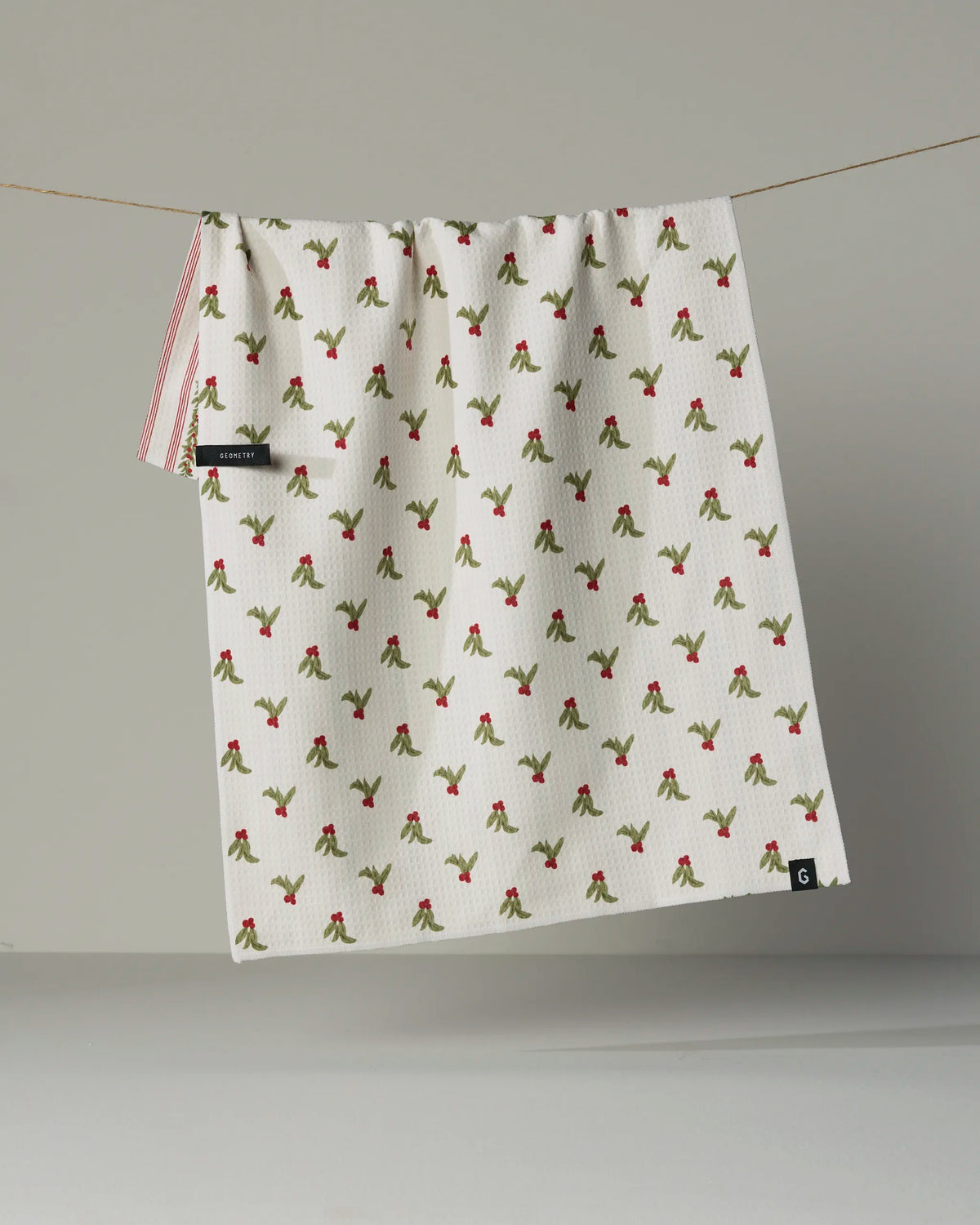 A white textured double-sided tea towel called “Holly Vine” by Geometry hangs from a line, featuring a vibrant repeating red and green cherry pattern. Made from recycled materials, it includes a small black tag and a corner label—blending eco-conscious practicality with stylish design.