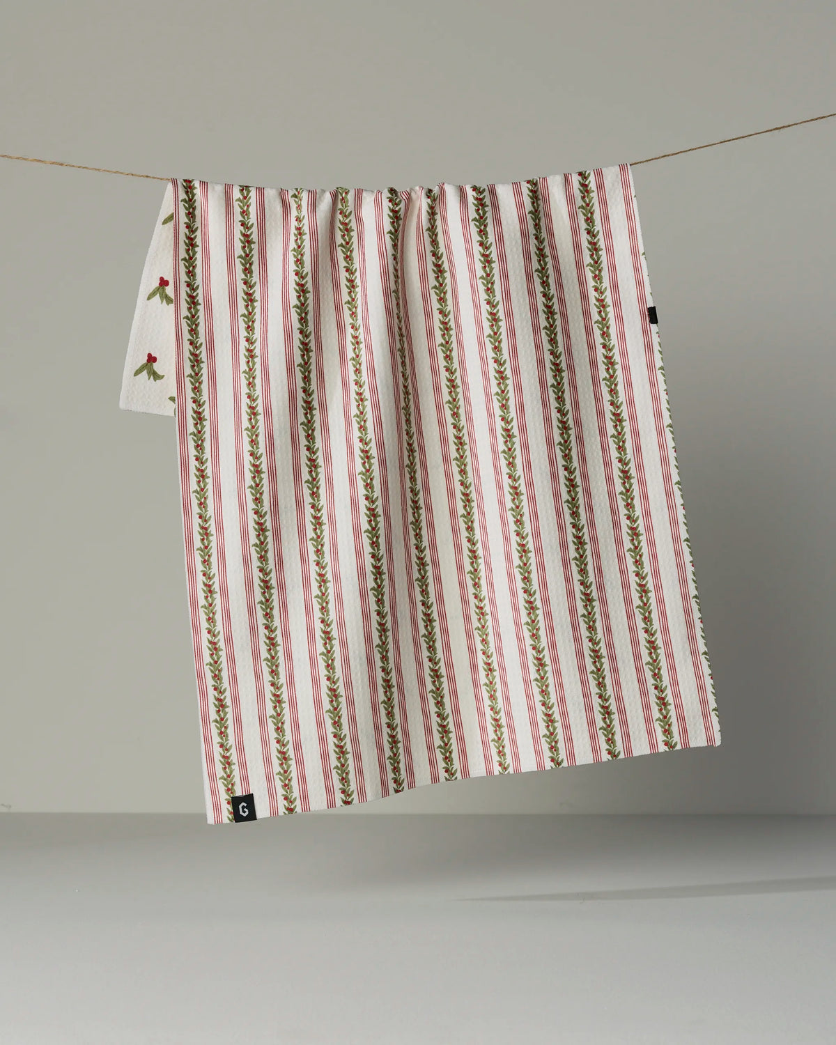 The Geometry “Holly Vine” tea towel, crafted from recycled materials, features red, green, and white vertical stripes with a small floral design detail. It is double-sided and hangs on a thin string against a plain gray background.