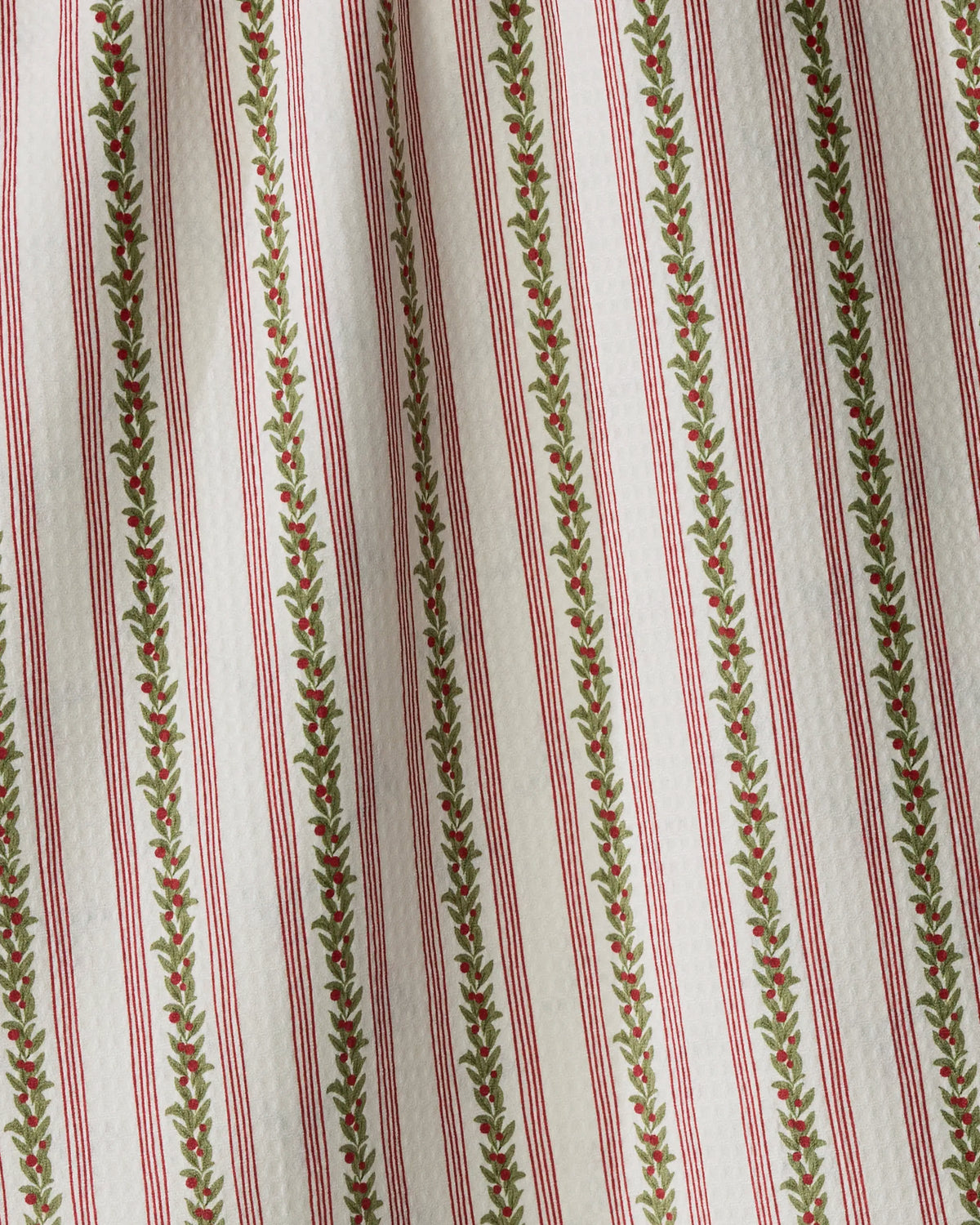 Close-up of Geometry’s Holly Vine fabric, featuring vertical red stripes and green leafy patterns on a white background. Crafted from recycled materials, this quick-drying textile displays slight folds that add shadow and texture, highlighting its quality and sustainability.