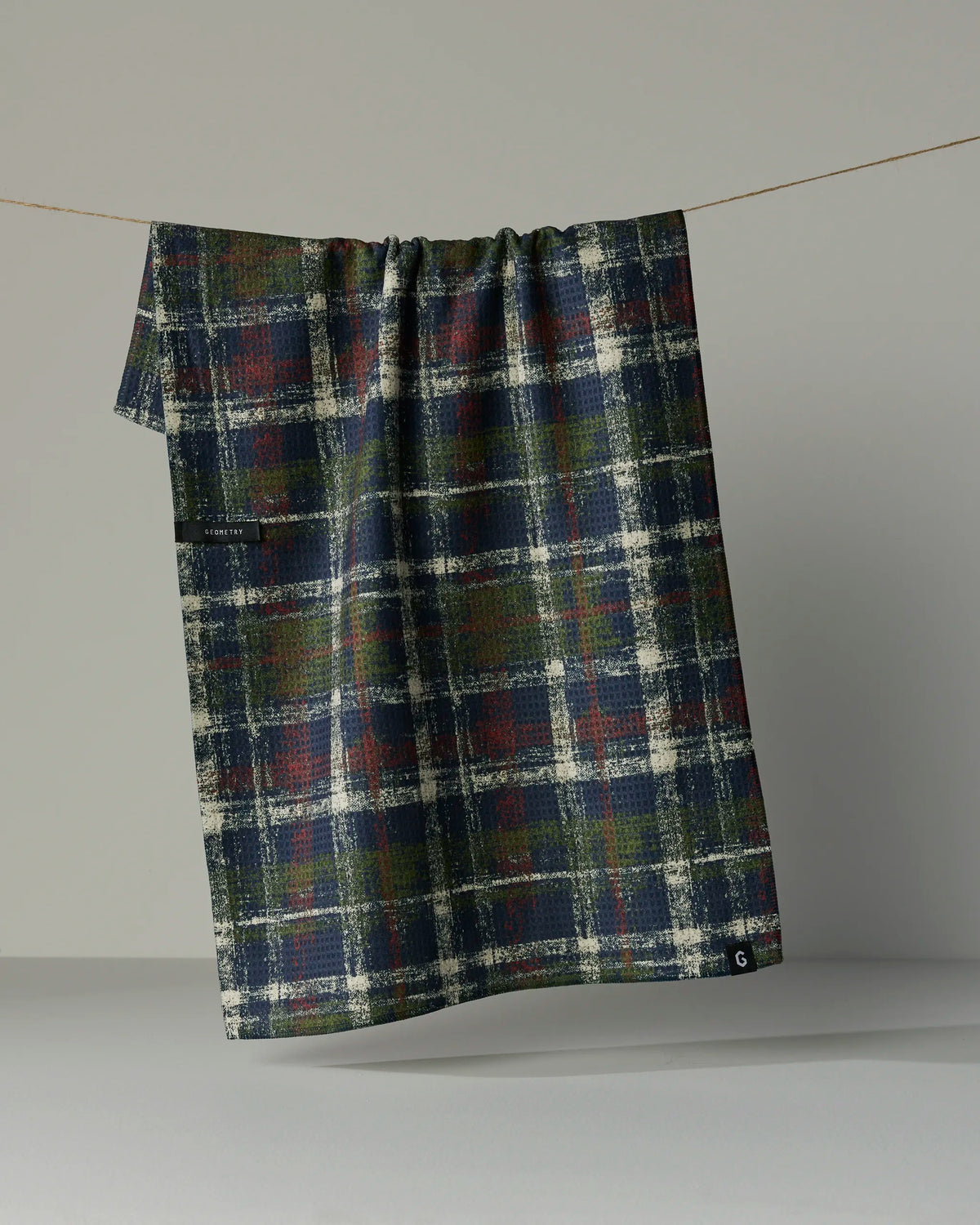 A Homestead Plaid double-sided tea towel by Geometry, featuring blue, green, red, and white plaid patterns, hangs neatly over a string against a plain light gray background.