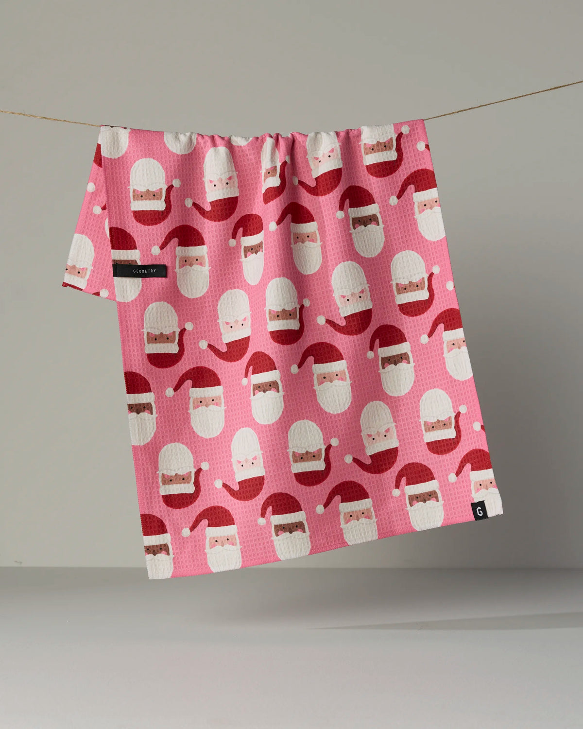 The “Jolly Santas” tea towel by Geometry features a vibrant pink design with a playful, repeating pattern of smiling Santa Claus faces in red hats. This quick-drying towel is shown hanging on a line against a plain background.