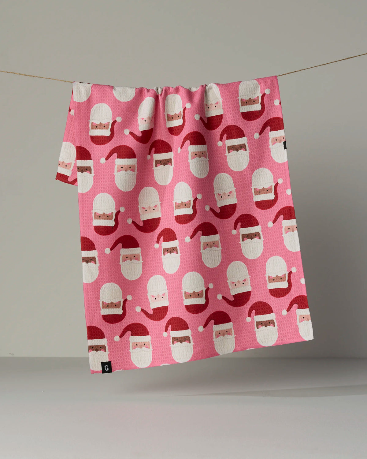 A “Jolly Santas” tea towel by Geometry, featuring a playful pink double-sided Santa Claus face pattern, hangs on a line against a neutral background.
