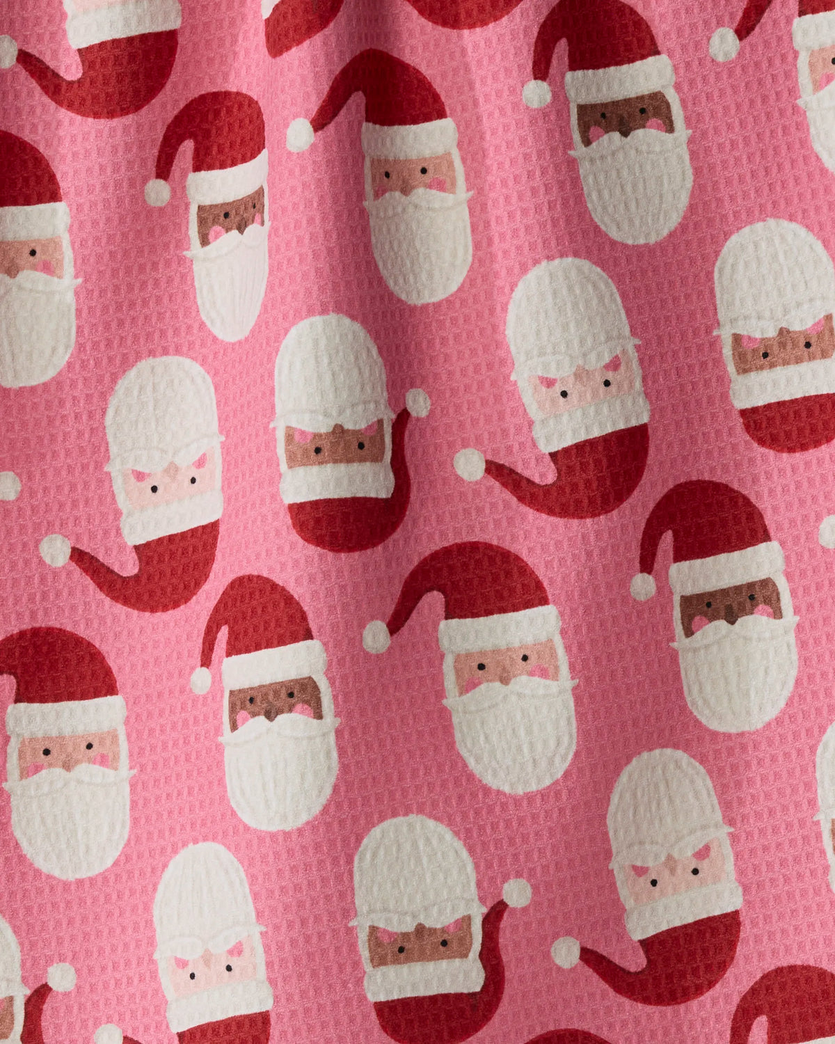 Jolly Santas by Geometry features a patterned fabric with cartoon Santa faces—some with light skin, others with dark skin—set against a pink background. The Santas sport white beards, red hats, and rosy cheeks. Crafted from recycled kitchen towel material, this fabric boasts a textured, waffle-like surface ideal for stylish and eco-friendly holiday crafts.