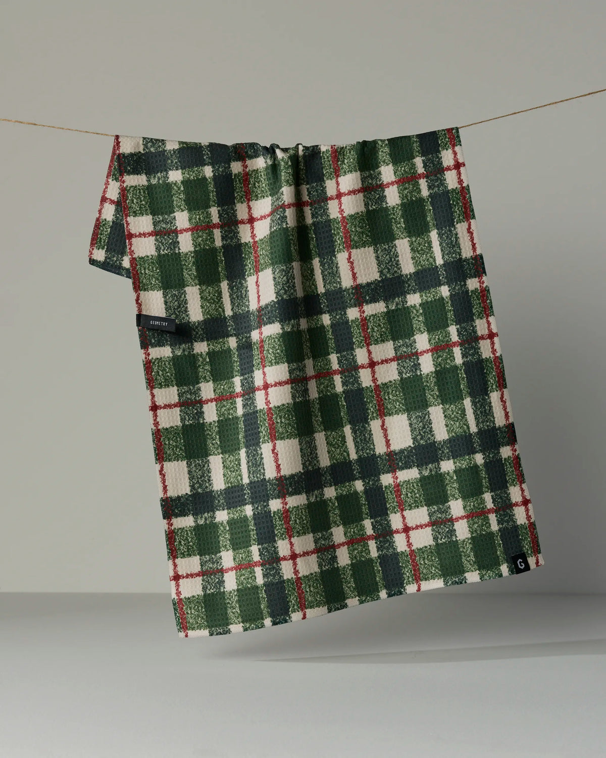 The Juniper Plaid double-sided tea towel by Geometry, featuring a green, red, and white plaid pattern, hangs on a thin line against a plain background, highlighting its quick-drying design made from recycled materials.