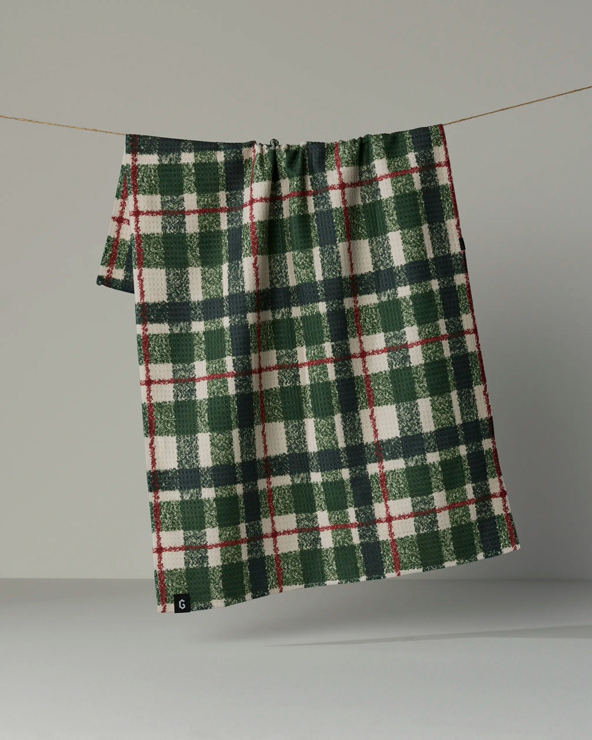 The Juniper Plaid by Geometry, a green, white, and red blanket made from recycled materials, hangs on a thin cord against a plain gray background.