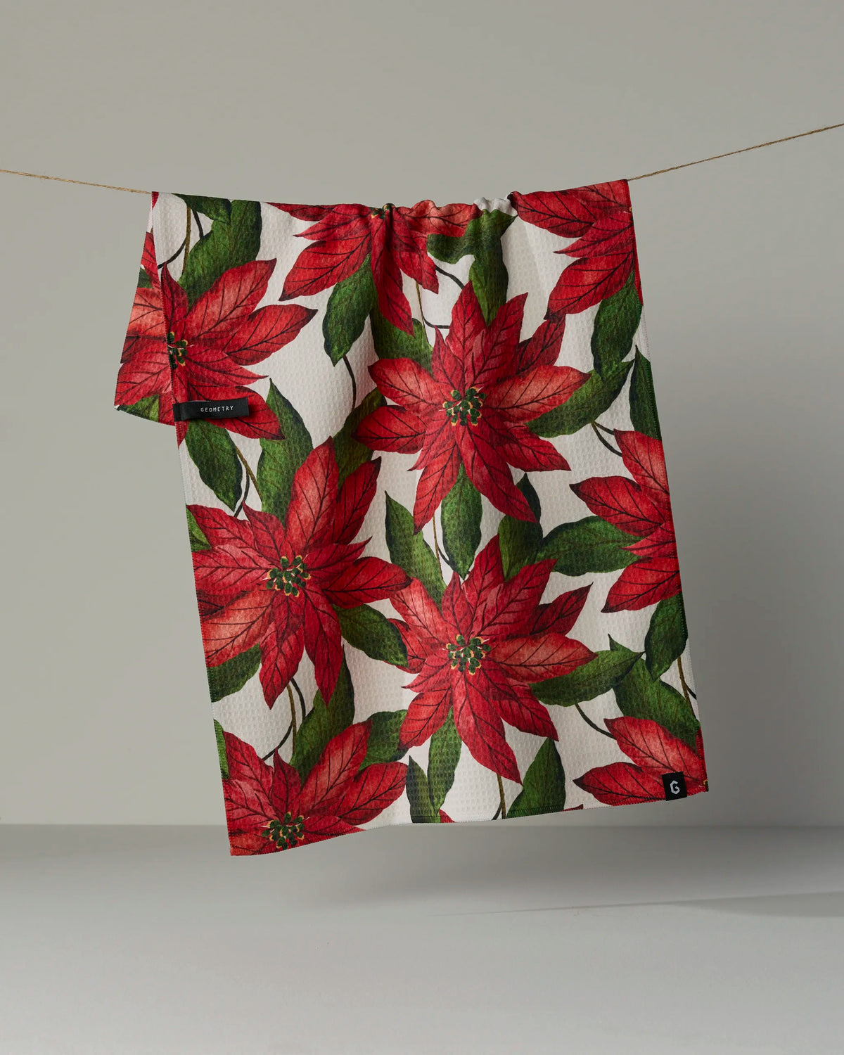 A quick-drying Geometry “Lauri” tea towel, featuring a vibrant red poinsettia and green leaves pattern, hangs on a thin string against a plain background.