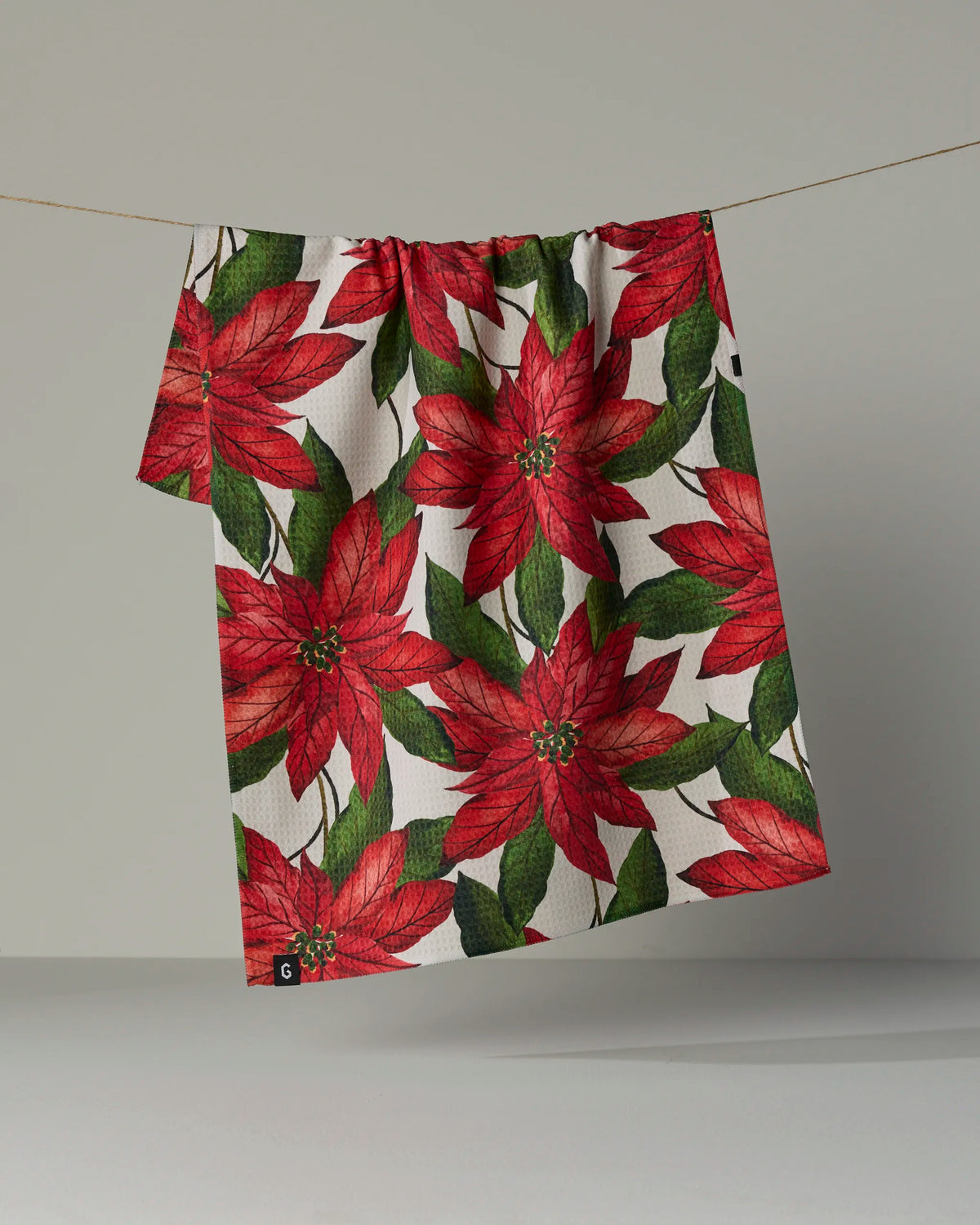 A quick-drying Geometry Lauri tea towel, featuring a red poinsettia and green leaf pattern, hangs on a line against a plain, neutral background.