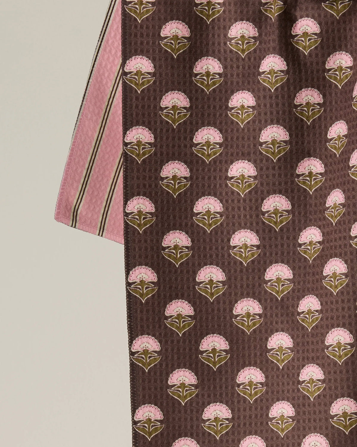 Introducing the Maisy tea towel from Geometry—a quick-drying towel that showcases a repeating pattern of pink flowers with green leaves set against a brown background, paired alongside another fabric featuring vertical pink and brown stripes.