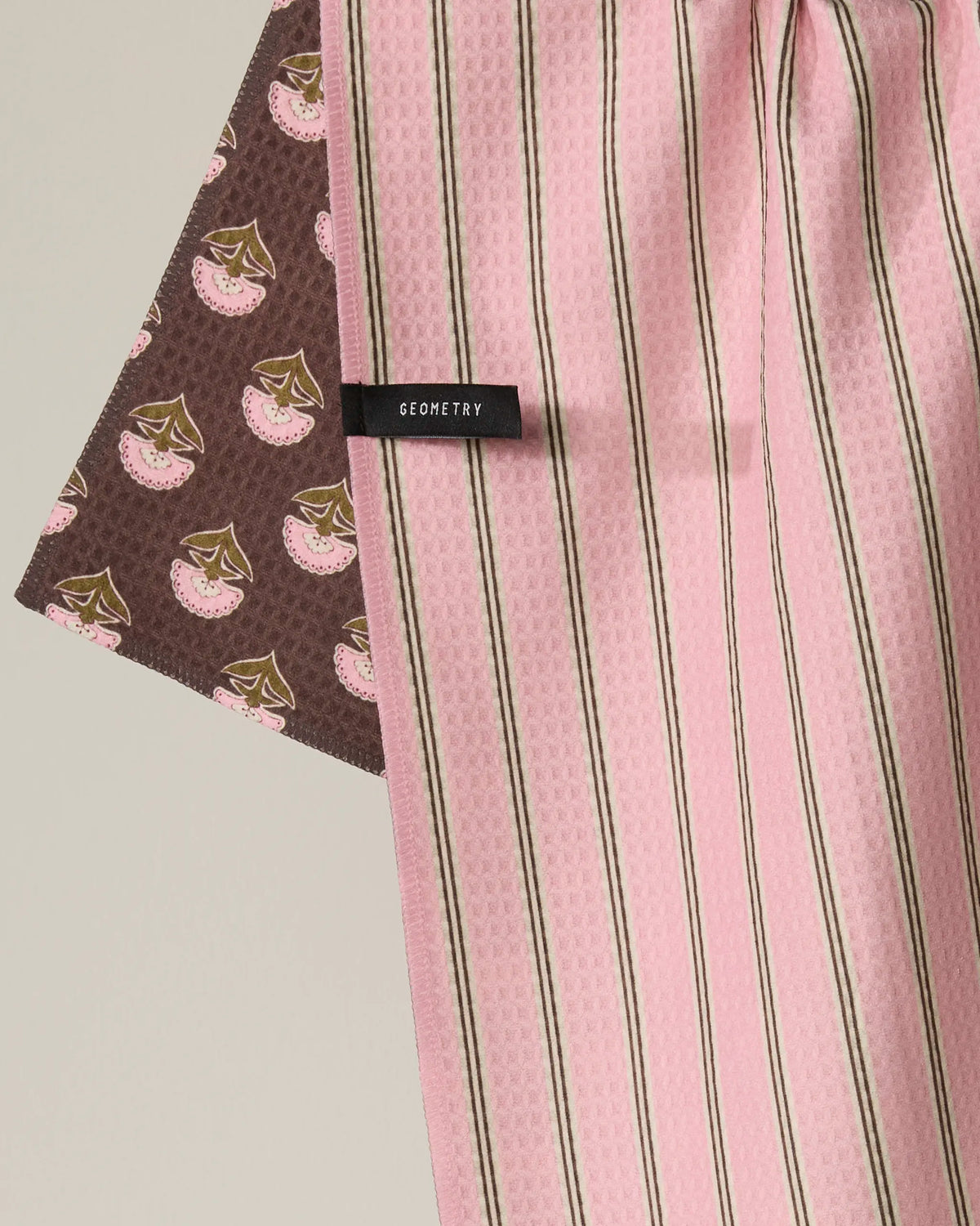 A close-up of Geometry’s Maisy double-sided tea towel, which features pink and brown vertical stripes on one side and a brown background with a repeating cherry pattern on the other. A small black label reading “GEOMETRY” is sewn onto this recycled kitchen towel.