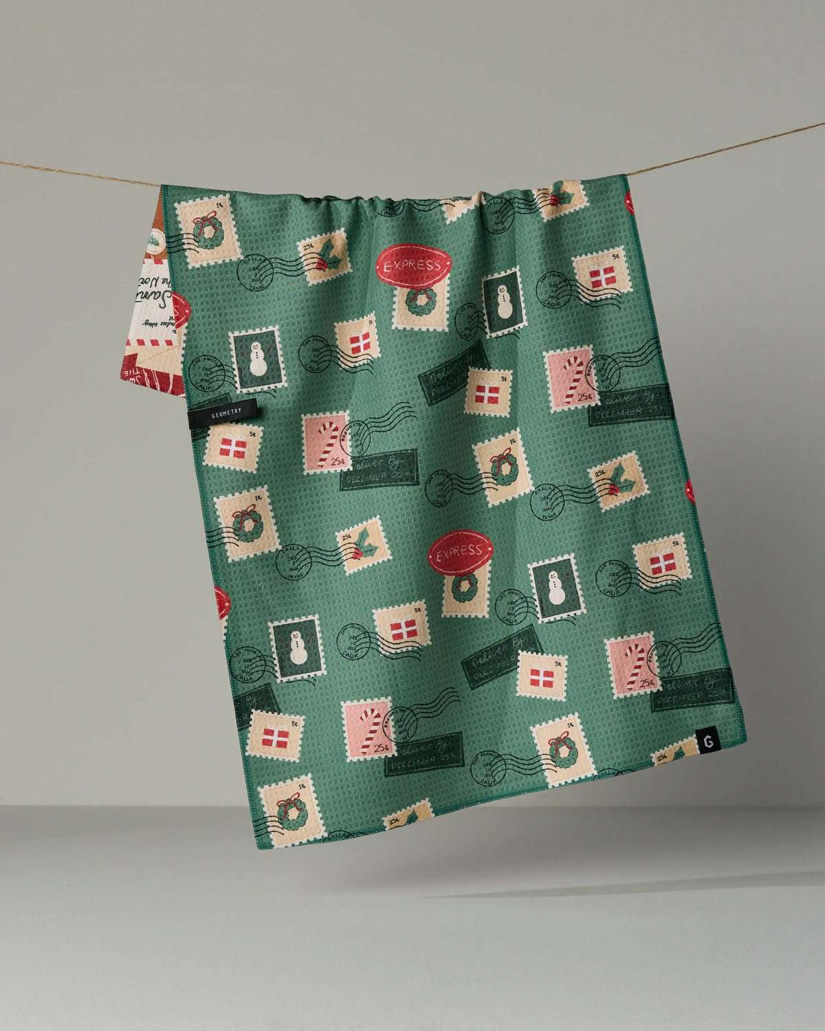 The Geometry Merry Mail tea towel, featuring a vibrant green background with a festive pattern of postage stamps, envelopes, wreaths, and postmarks in shades of red, white, pink, and green, adds holiday charm to your kitchen. This quick-drying towel is perfect for keeping up with busy holiday preparations.