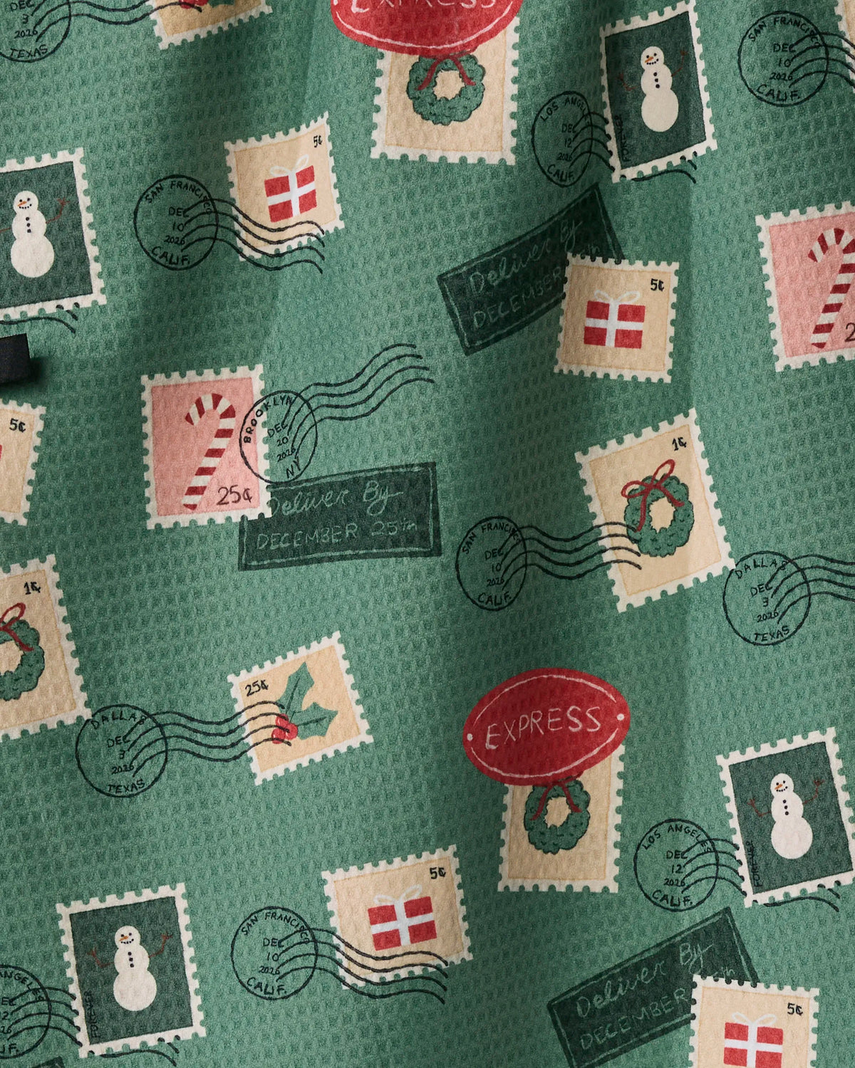 The “Merry Mail” double-sided tea towel from Geometry features green fabric decorated with holiday-themed postage stamp patterns—including candy canes, wreaths, snowmen, and gift boxes—alongside festive postmarks and labels like “EXPRESS” and “Deliver By December 25th.” This cheerful towel is both festive and functional, making it the perfect way to spread holiday cheer in your kitchen!.