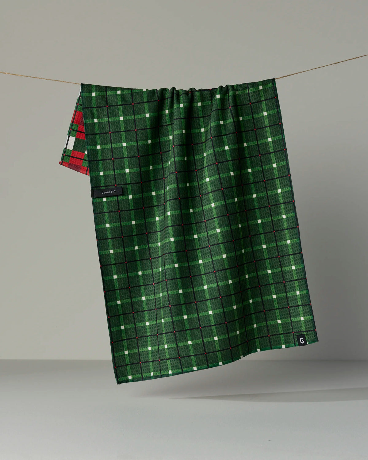 A green Noel Plaid tea towel by Geometry, featuring black and white lines with a touch of red plaid at the corner, hangs on a line. Crafted from recycled materials, this double-sided kitchen towel combines style and sustainability for your home.