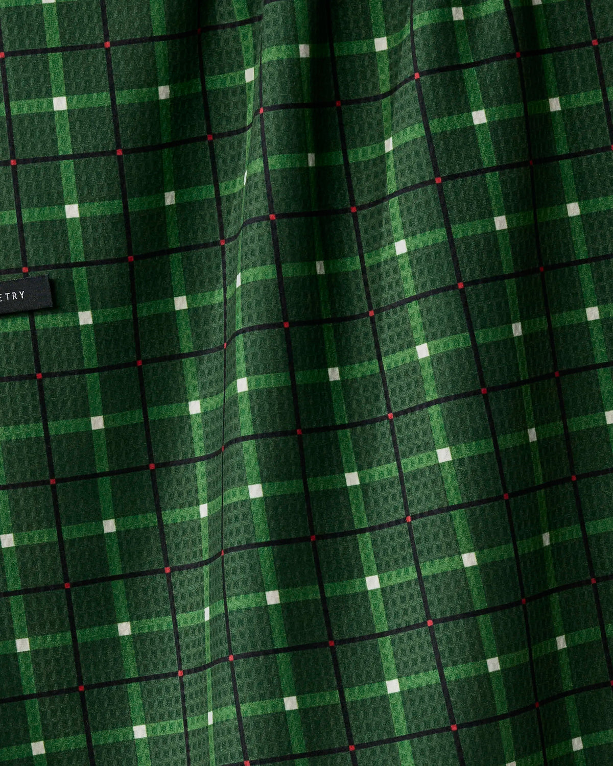 Close-up of Geometry’s Noel Plaid fabric, showcasing a green material with a black grid pattern and small light green squares at the intersections; features a subtle texture crafted from recycled materials and includes a black fabric tag on the left.