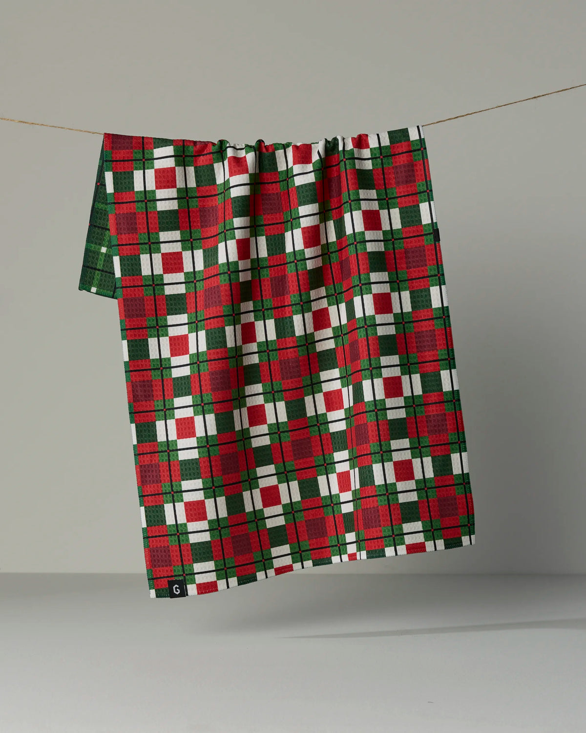 The Noel Plaid tea towel by Geometry, featuring a double-sided red, green, and white plaid design, hangs on a string against a plain gray background. Crafted from recycled materials, this quick-drying cloth offers both style and sustainability.