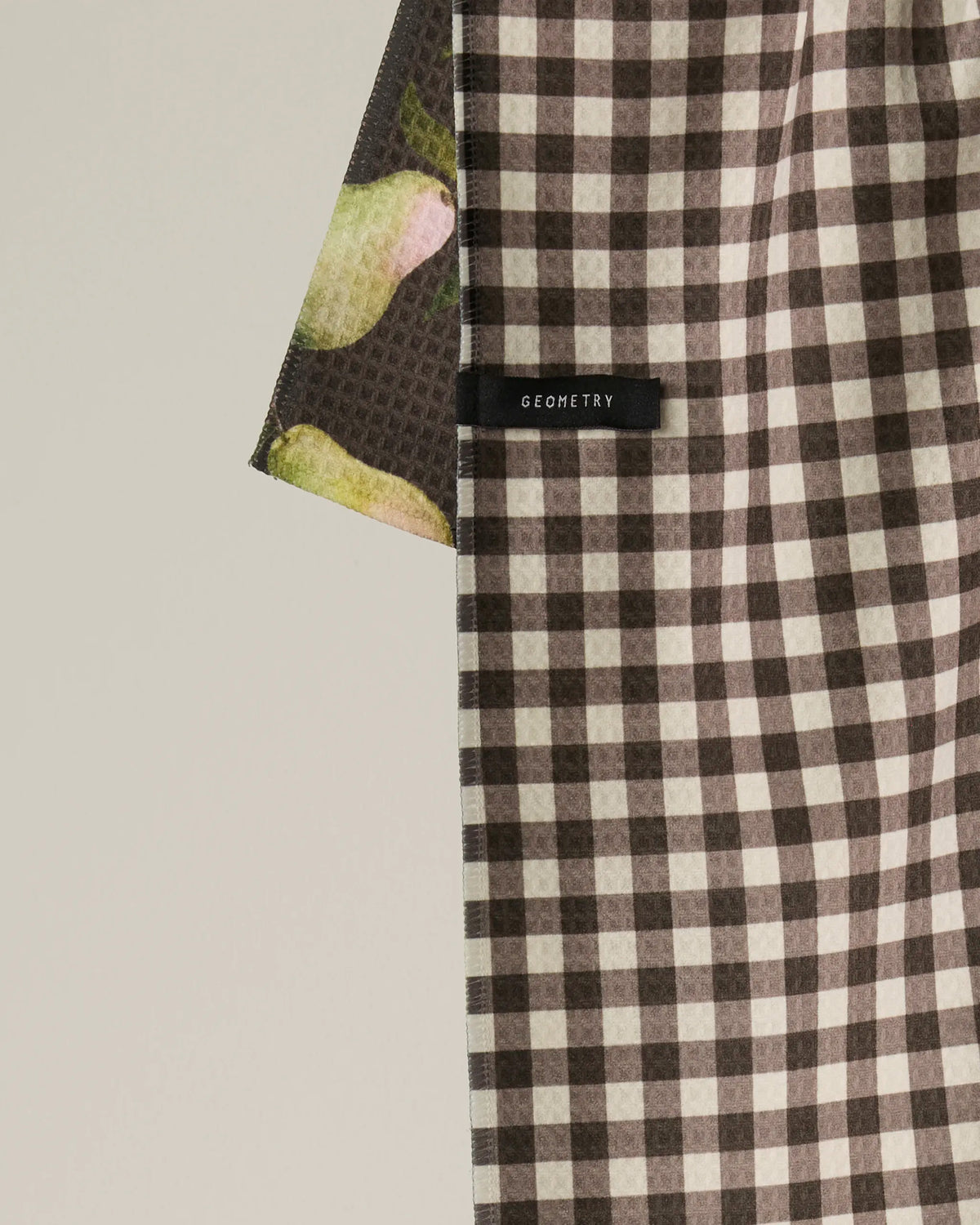 A close-up of Geometry’s Pearwood towel in a black and white checkered pattern features a small black tag labeled GEOMETRY, while another towel with a green and pink design made from recycled materials slightly overlaps on the left side.