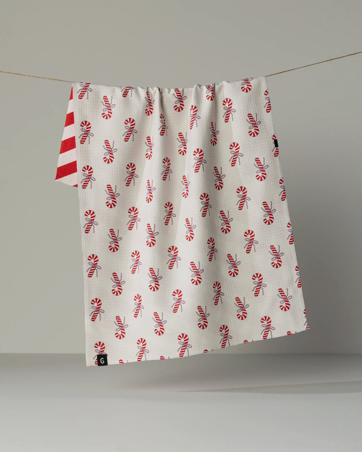 Geometry’s Peppermint Lane double-sided tea towel features playful red and white peppermint candies and festive bow patterns, all bordered by vibrant red and white stripes. Crafted from Geometry’s signature quick-drying fabric, this towel is ideal for daily use. A softly lit background accentuates the charming design as it hangs on a slender line.