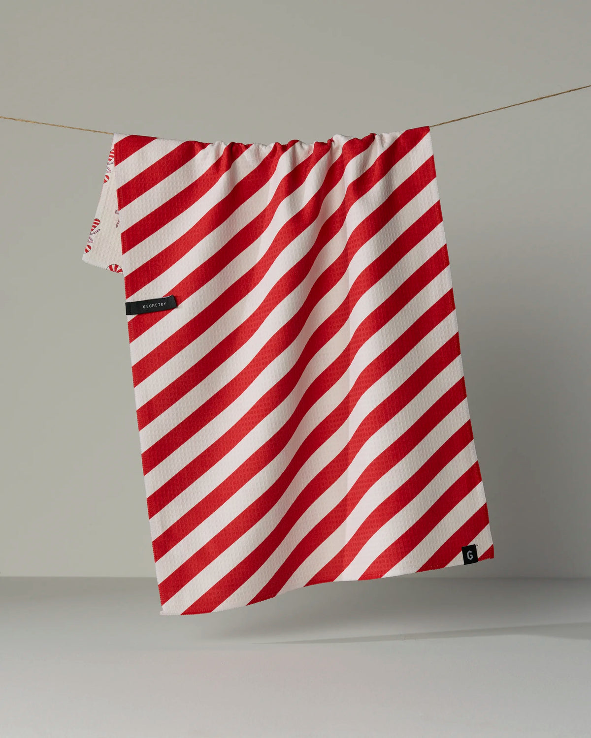 The Geometry Peppermint Lane double-sided tea towel, featuring red and white diagonal stripes and a small black label on one corner, hangs on a clothesline against a plain gray background.