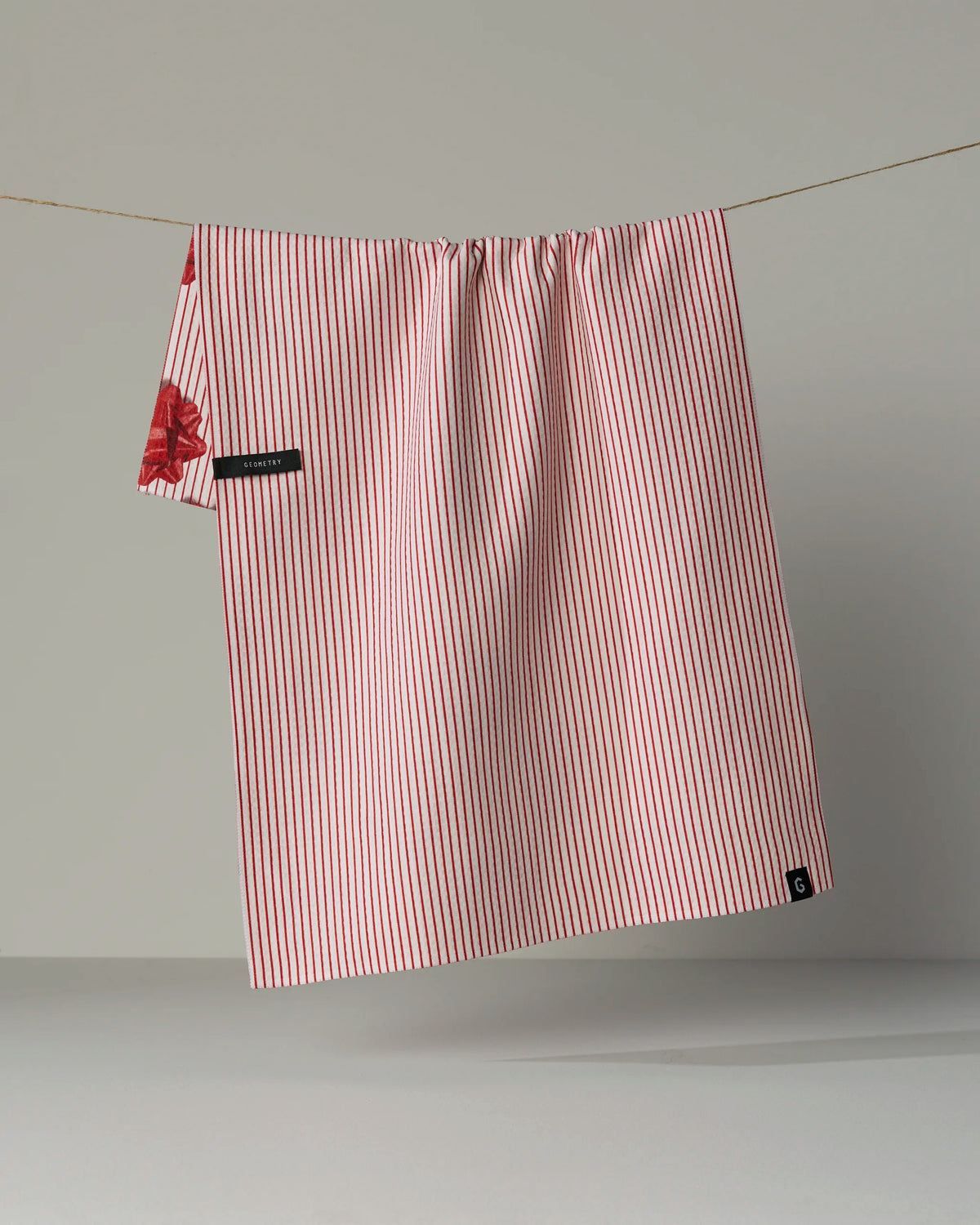 The Geometry “Perfectly Wrapped” double-sided tea towel, displaying red and white stripes with a touch of floral pattern, hangs on a clothesline against a plain background.