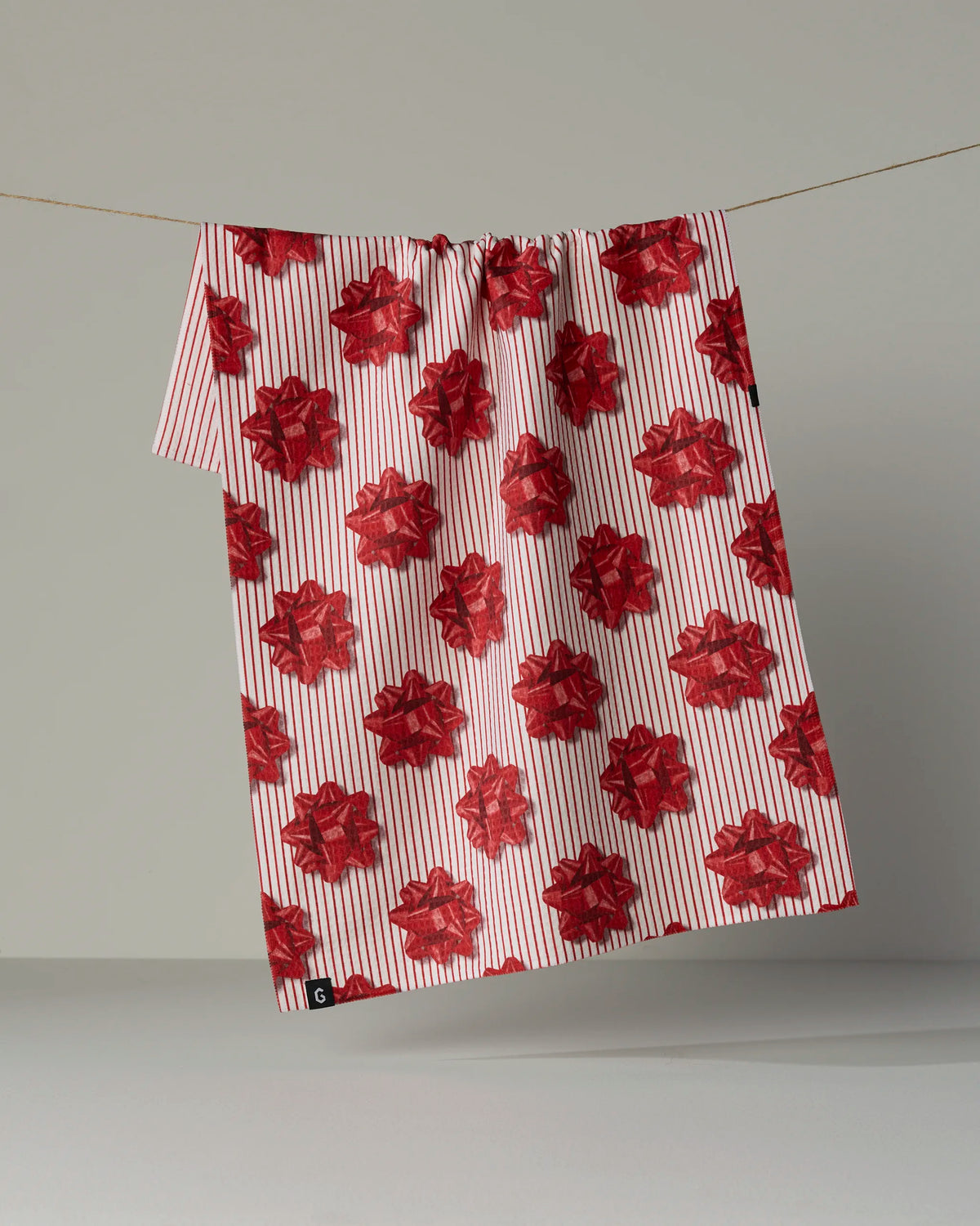 The “Perfectly Wrapped” tea towel by Geometry, featuring red gift bow patterns and thin red vertical stripes on both sides, hangs on a line against a plain gray background.