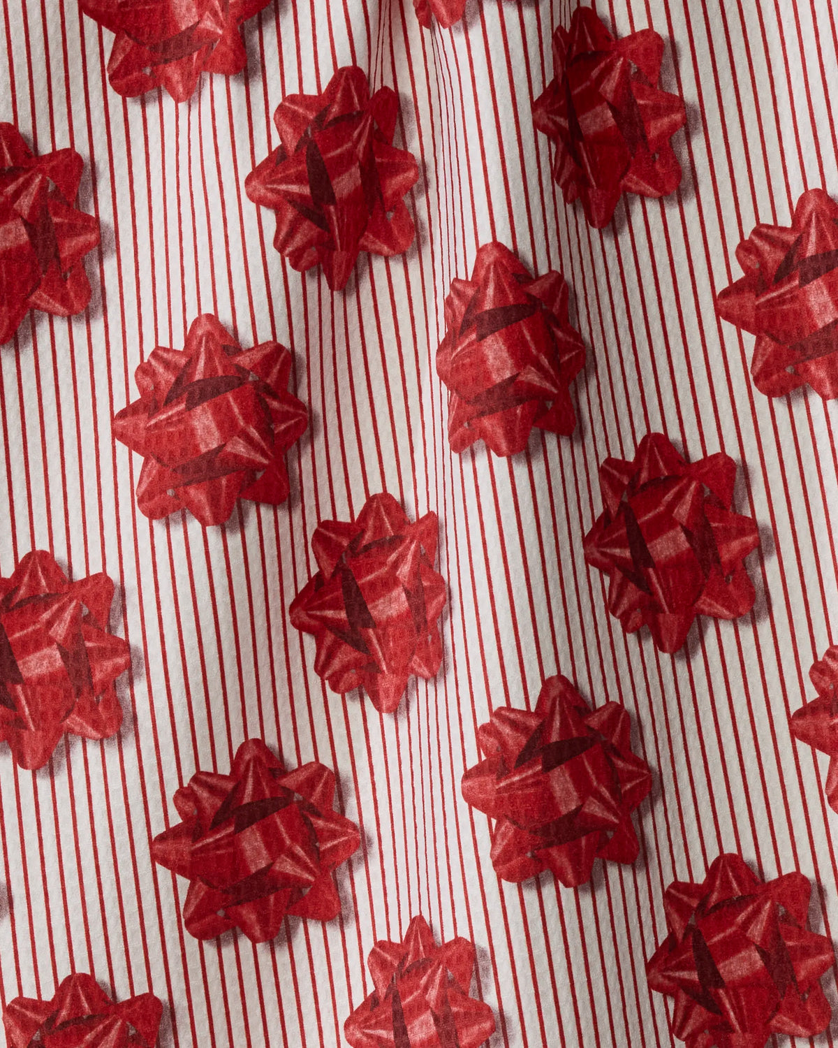 Red gift bows adorn white wrapping paper accented with vertical red stripes, their design brought to life by subtle folds that add depth and texture. The repeating bow pattern recalls the festive look of the “Perfectly Wrapped” double-sided tea towel from Geometry—an accessory sure to add charm to any kitchen décor.
