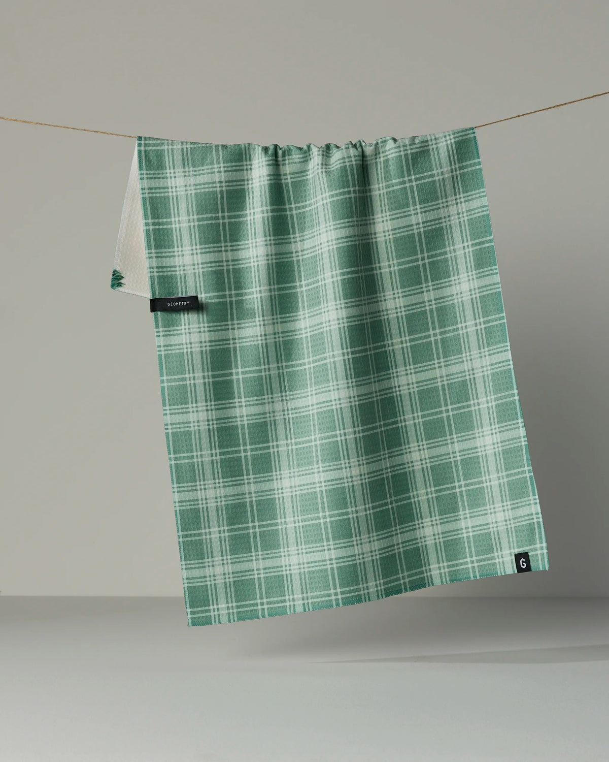 The Pine Grove tea towel by Geometry, featuring a green and white plaid pattern, hangs on a string against a plain, light background. Absorbent and quick-drying, this recycled kitchen towel includes a small black tag on one side.