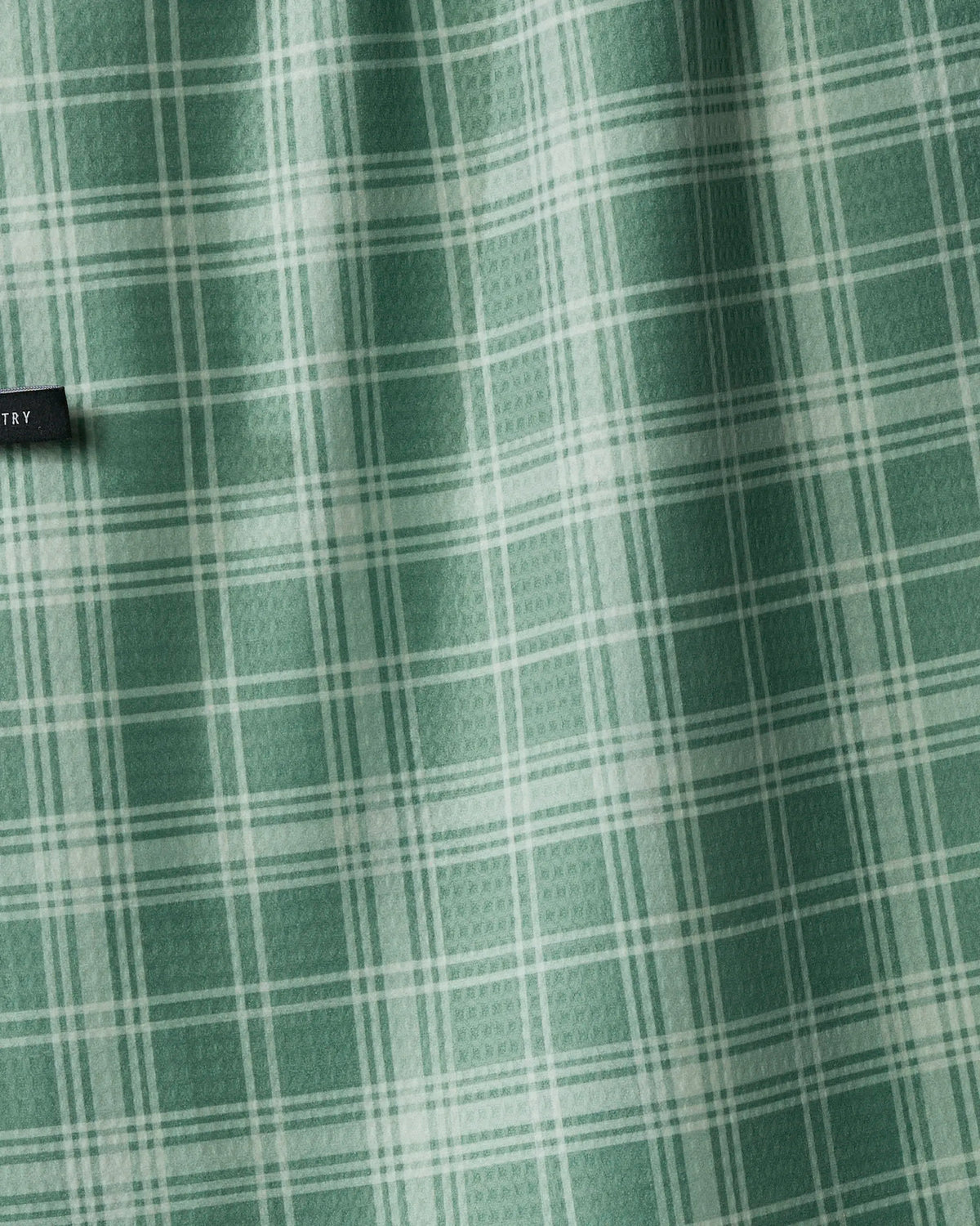 The Geometry Pine Grove tea towel features a green and white plaid pattern on smooth, absorbent quick-dry fabric—ideal for an eco-friendly kitchen. Subtle light and shadow reveal gentle folds in the recycled material, with a small black Geometry tag visible at the upper left edge.