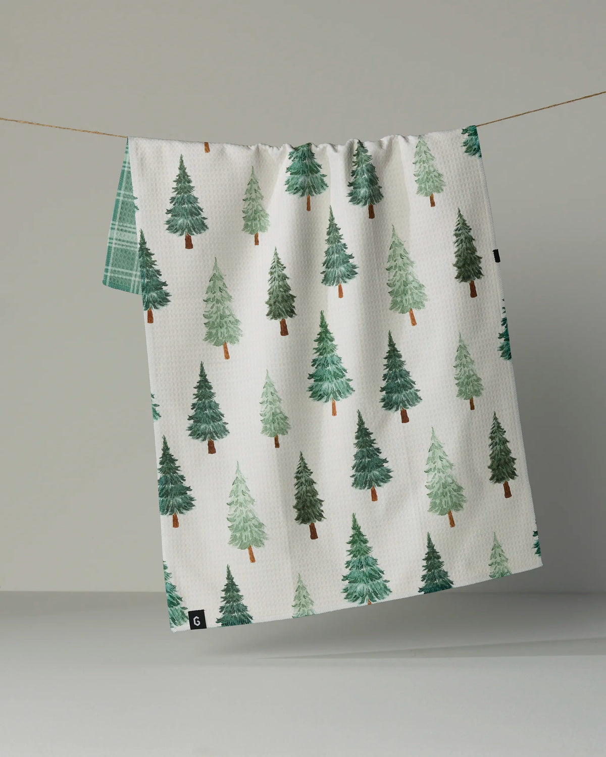 A Geometry “Pine Grove” recycled kitchen towel, featuring a pattern of green pine trees on white, hangs on a line against a plain gray background. A section of green plaid fabric can be seen behind this absorbent and quick-dry tea towel.