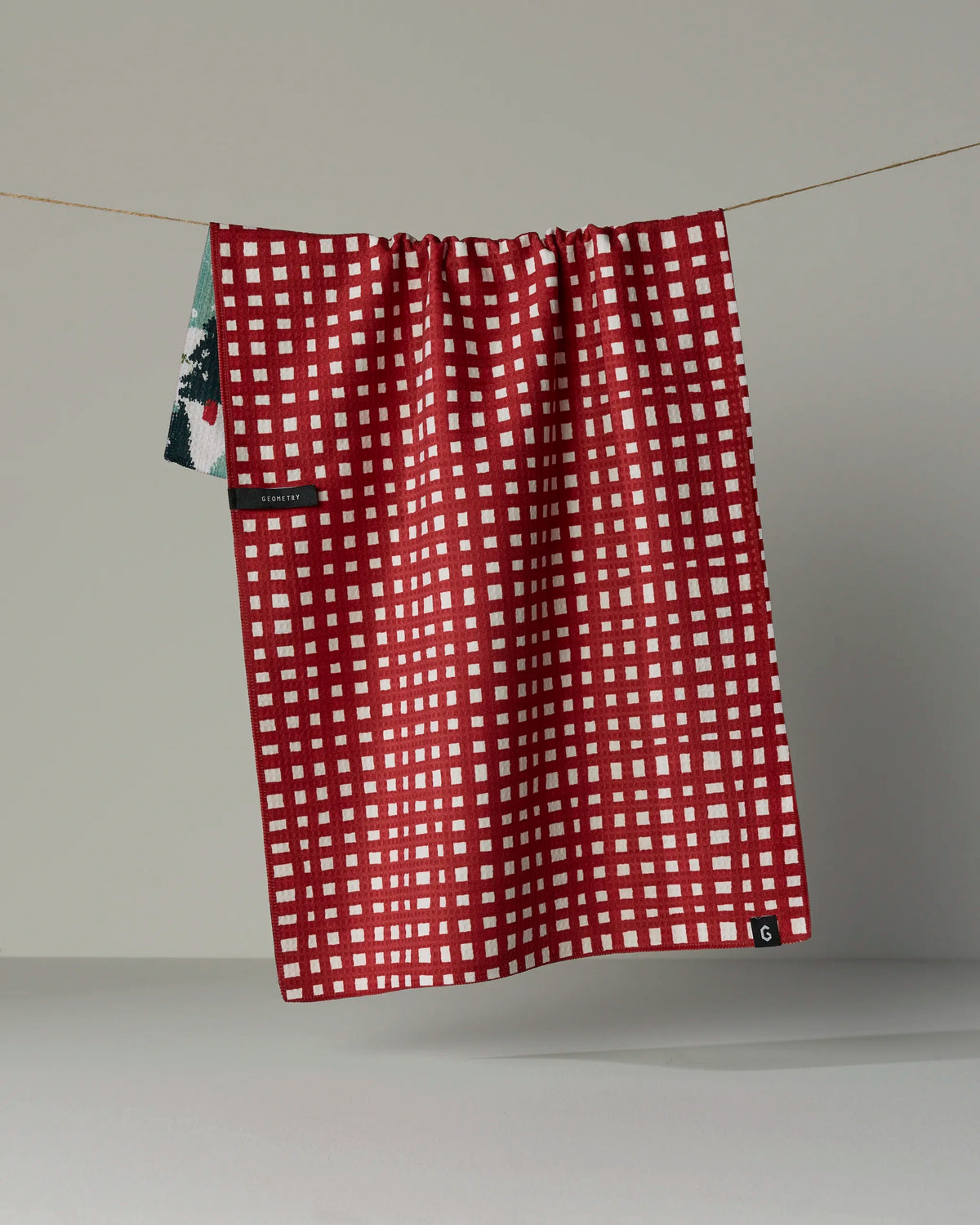 A red cloth with a white grid pattern, named “Pine Parade” by Geometry and crafted from recycled kitchen towel material, hangs on a string against a plain gray background. A small tag is visible on the lower right corner of the absorbent, quick-drying towel.
