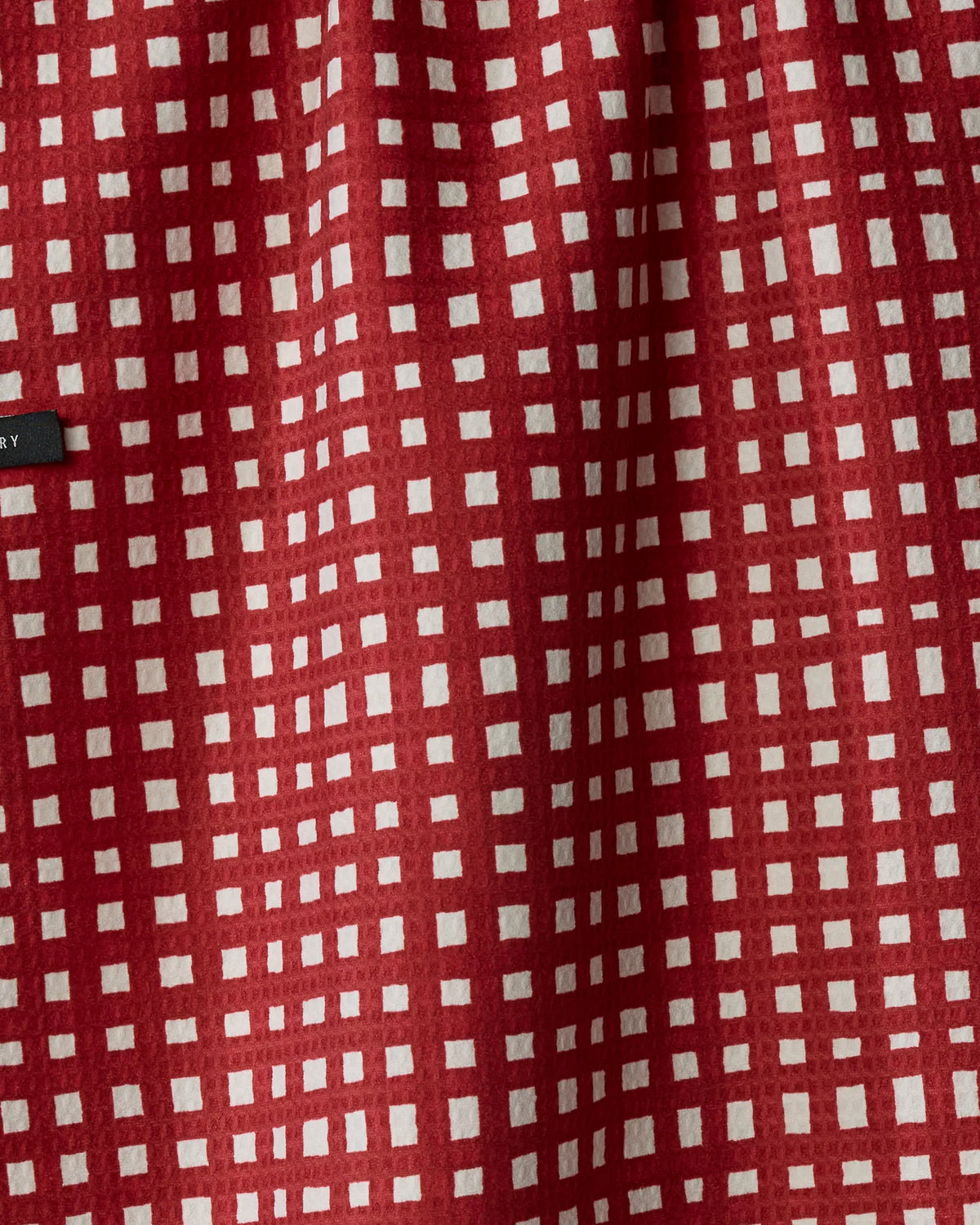 Crafted by Geometry, the “Pine Parade” tea towel features a red and white checkered design with irregular grid lines, creating a distinctive woven pattern. This double-sided towel is both absorbent and quick-drying—perfect for busy kitchens. A small black Geometry label is partially visible along the left edge of the fabric.