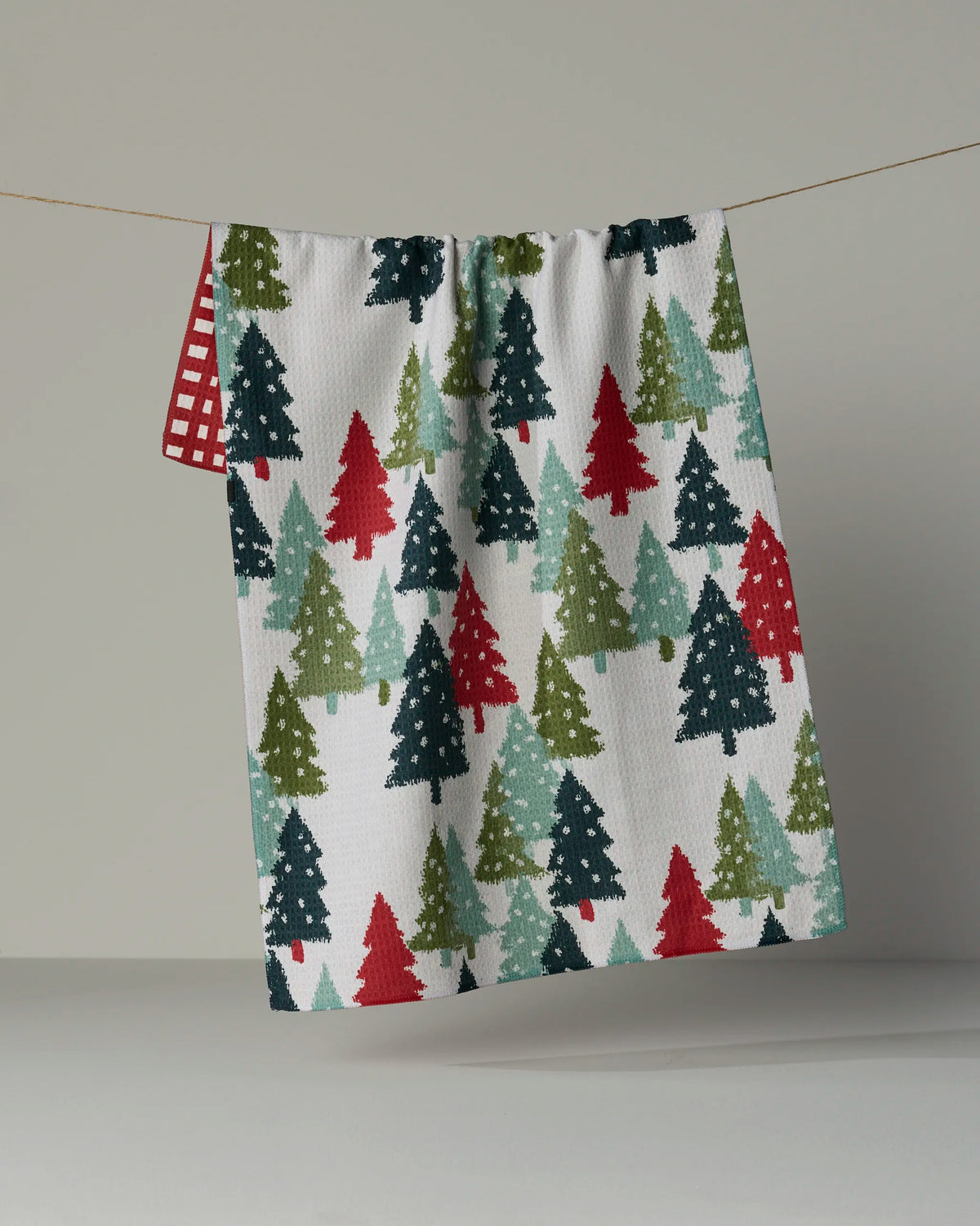 A festive double-sided tea towel from Geometry, called “Pine Parade,” featuring a pattern of green, red, and teal Christmas trees hangs on a string. Behind it, a second absorbent and quick-drying towel with a red and white checkered design is partially visible.